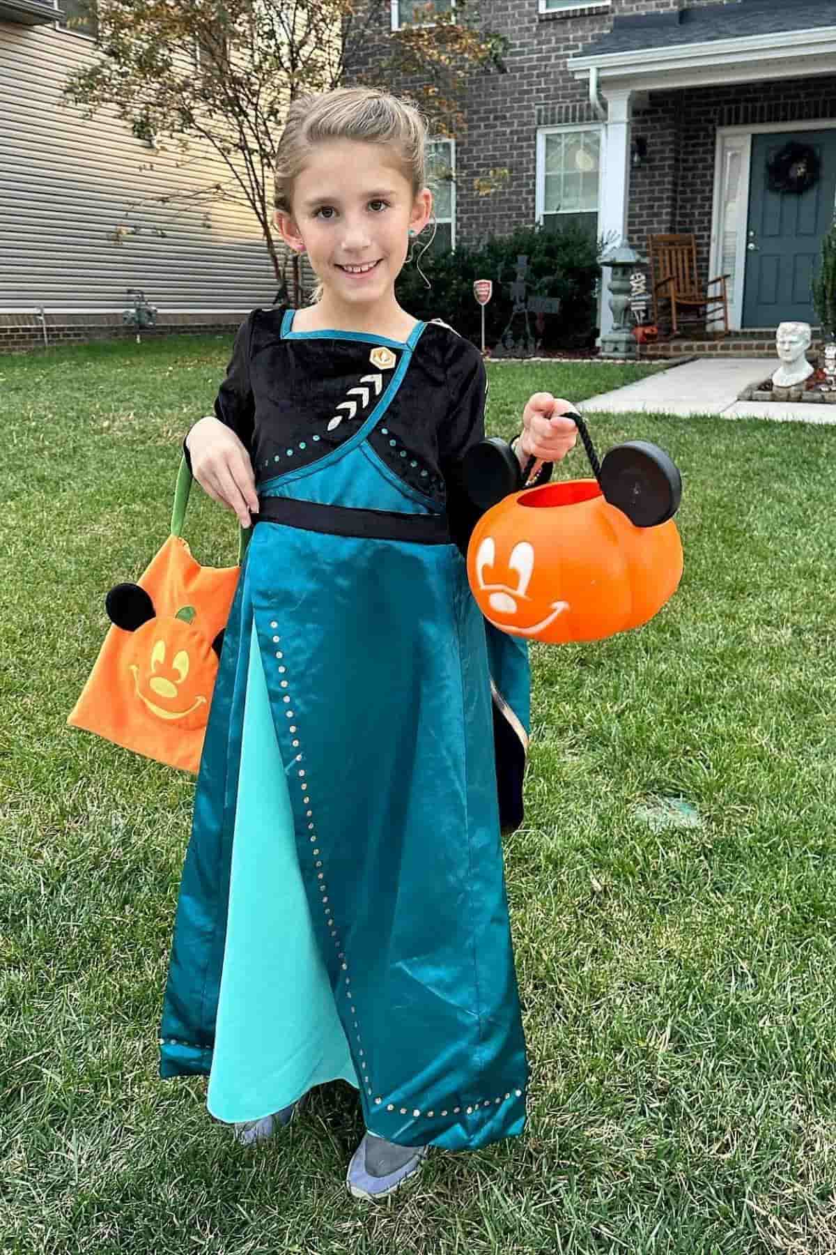 A young girl in a teal and black princess costume stands on grass, holding two orange pumpkin-shaped trick-or-treat buckets.