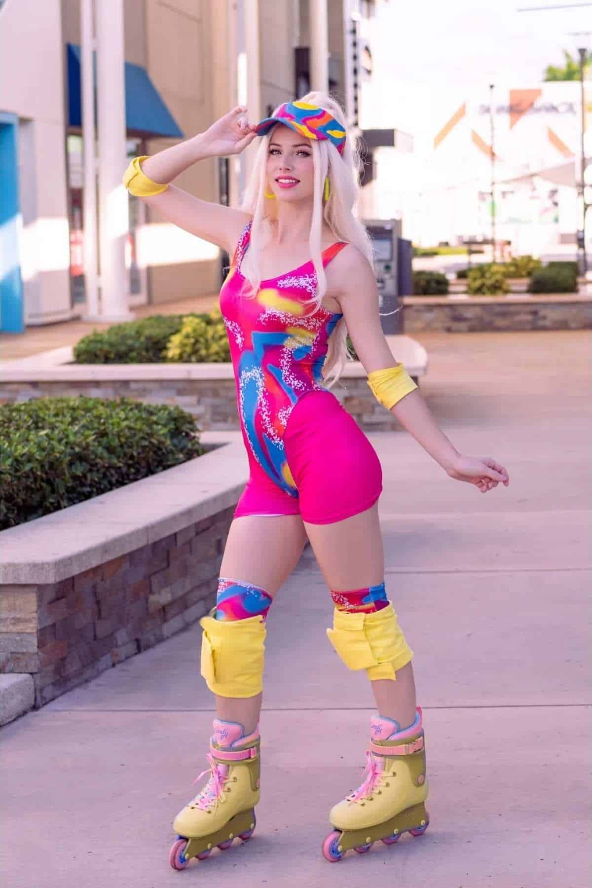 Person in a pink and blue roller skating outfit with yellow pads and a matching cap poses outdoors on a paved sidewalk.