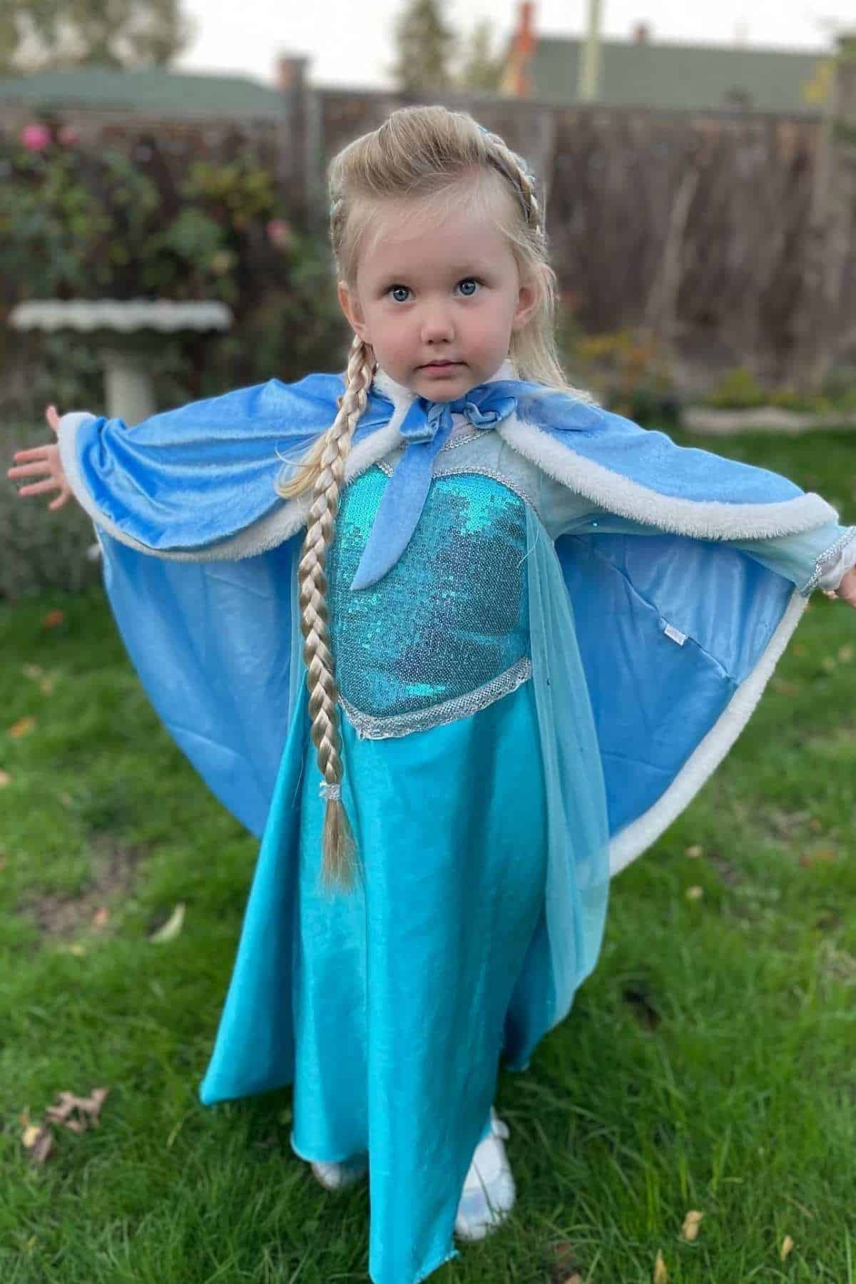 Young child in a blue and turquoise princess costume stands with arms outstretched on grass, wearing a long blonde braid and a blue cape in a backyard setting.
