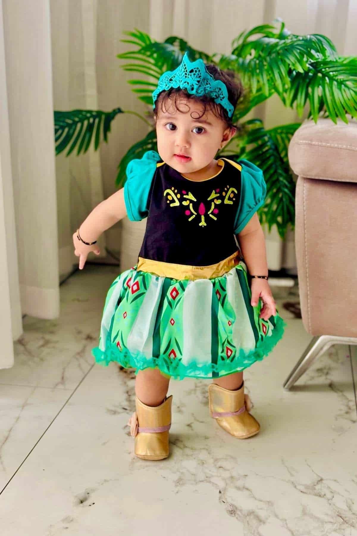 A toddler wearing a colorful dress, gold shoes, and a blue headband stands indoors on a marble floor near a sofa and green plants.