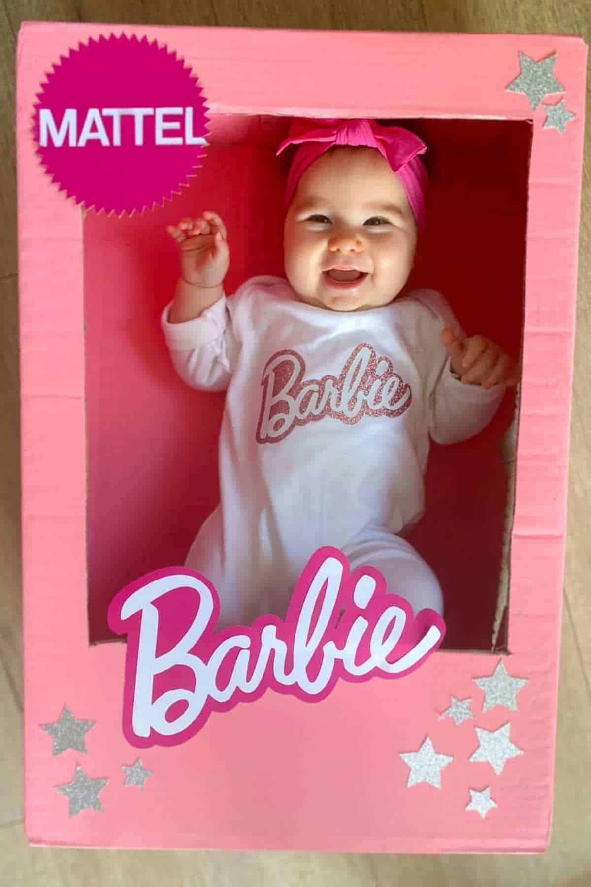 A smiling baby dressed in a Barbie-themed outfit sits in a pink cardboard box designed to look like Barbie doll packaging, with stars and logos.