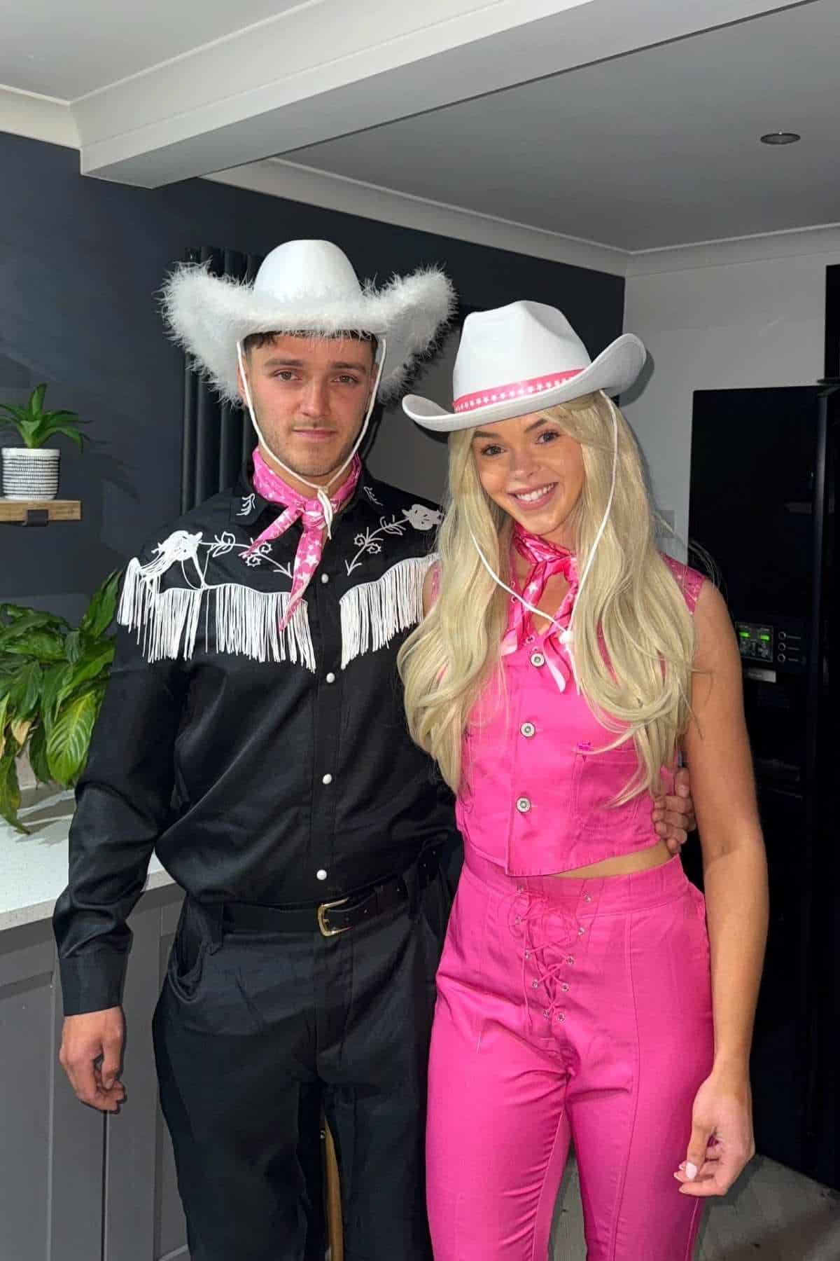 A man and woman wearing cowboy hats and western outfits stand together in a kitchen; he wears black, she wears pink.