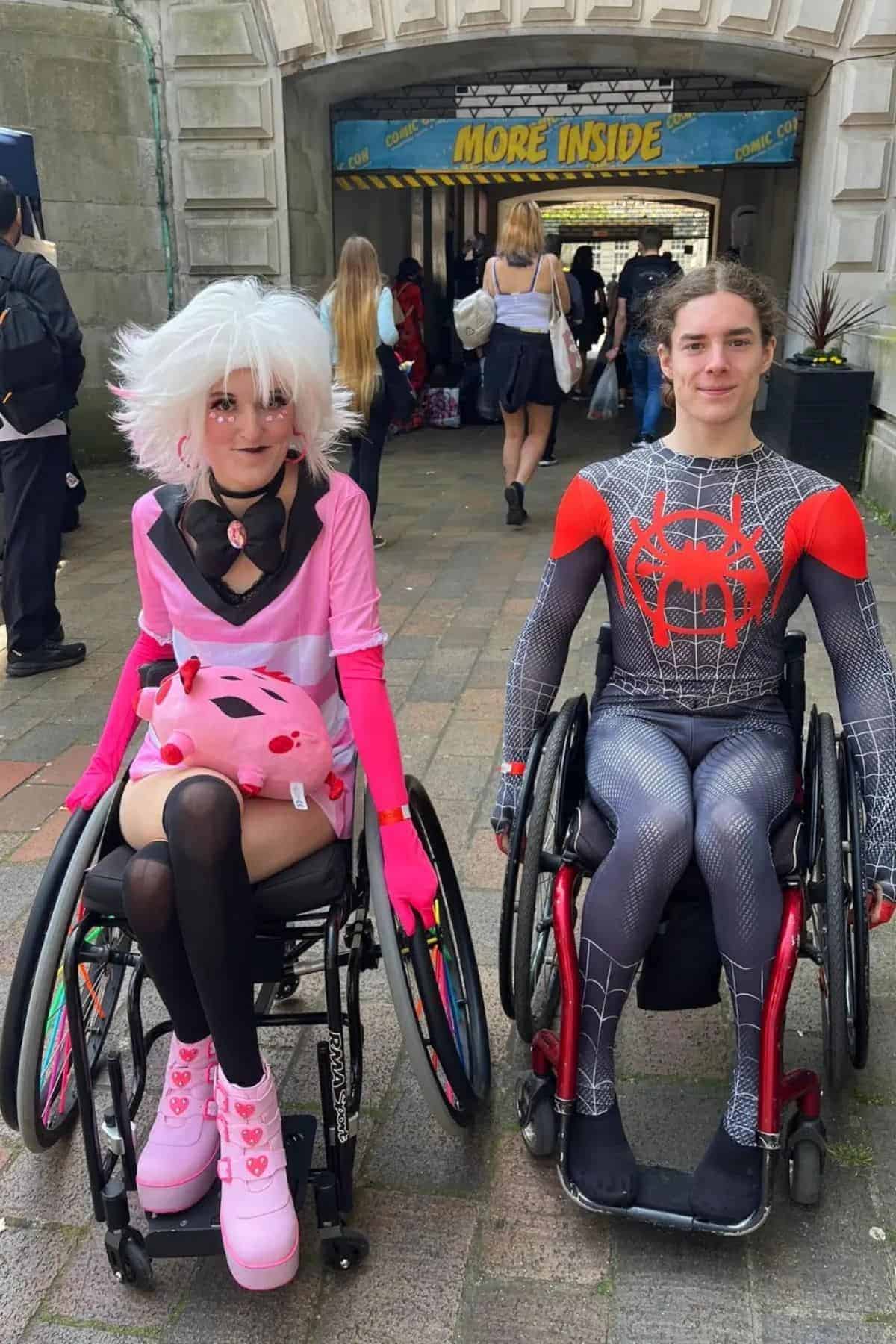 Two people in wheelchairs wearing cosplay costumes, sitting side by side outdoors at a convention entrance with others in the background.
