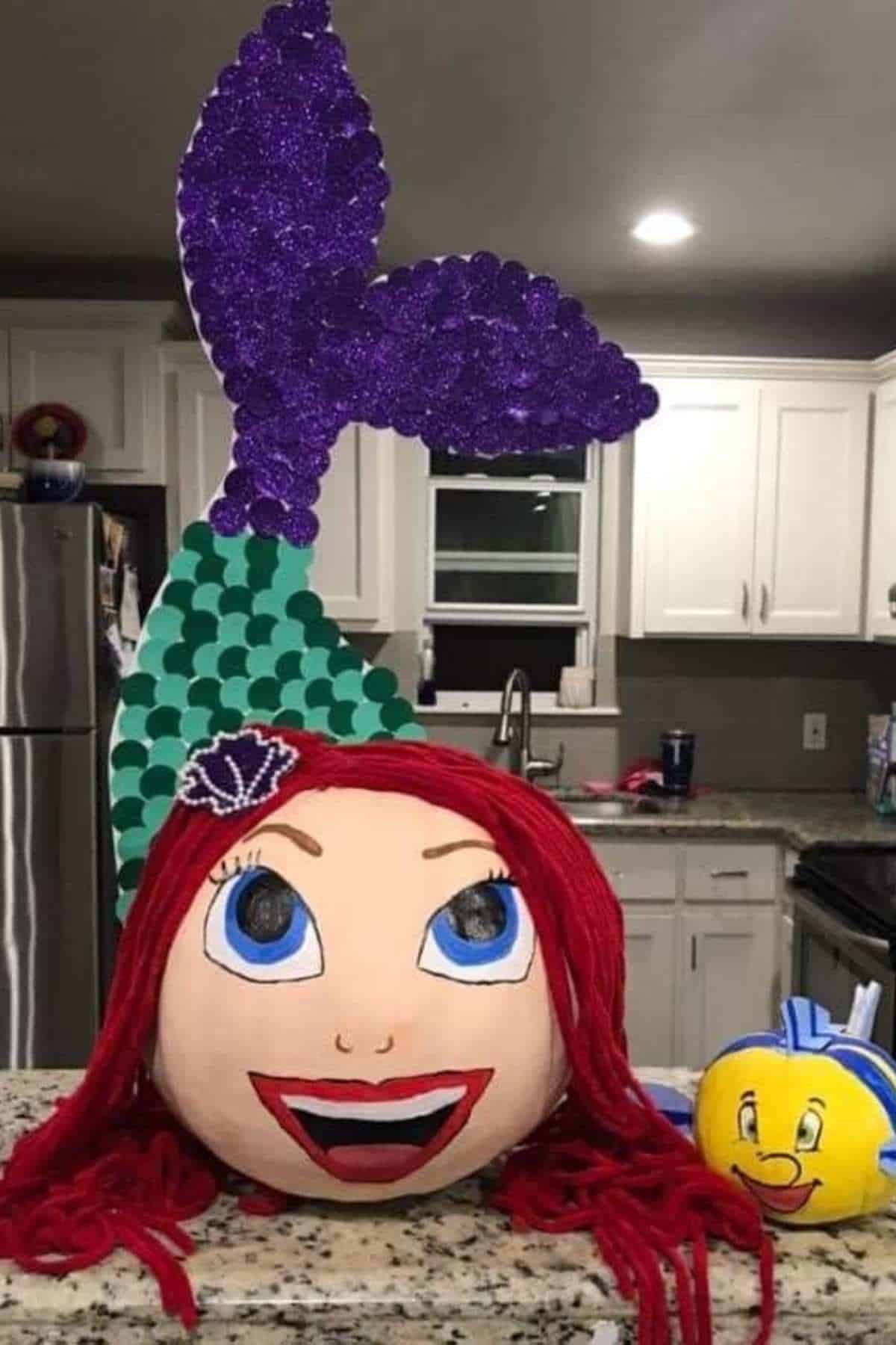 A pumpkin decorated as a mermaid with red yarn hair and a purple tail, next to a small yellow fish pumpkin, on a kitchen counter.