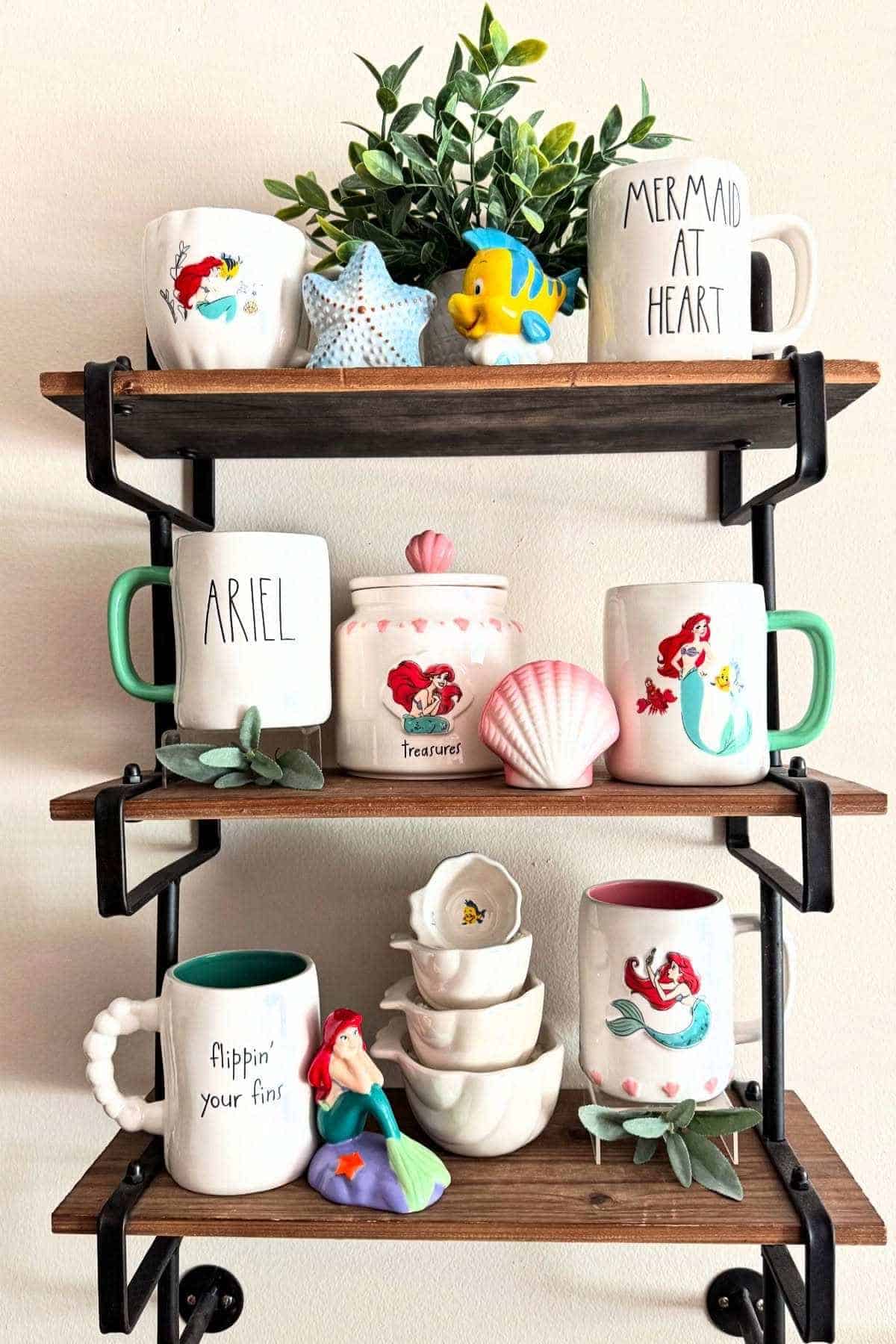Three wooden shelves display mugs, figurines, and small dishes, all themed around Ariel from The Little Mermaid, along with sea-inspired decor and a small green plant.