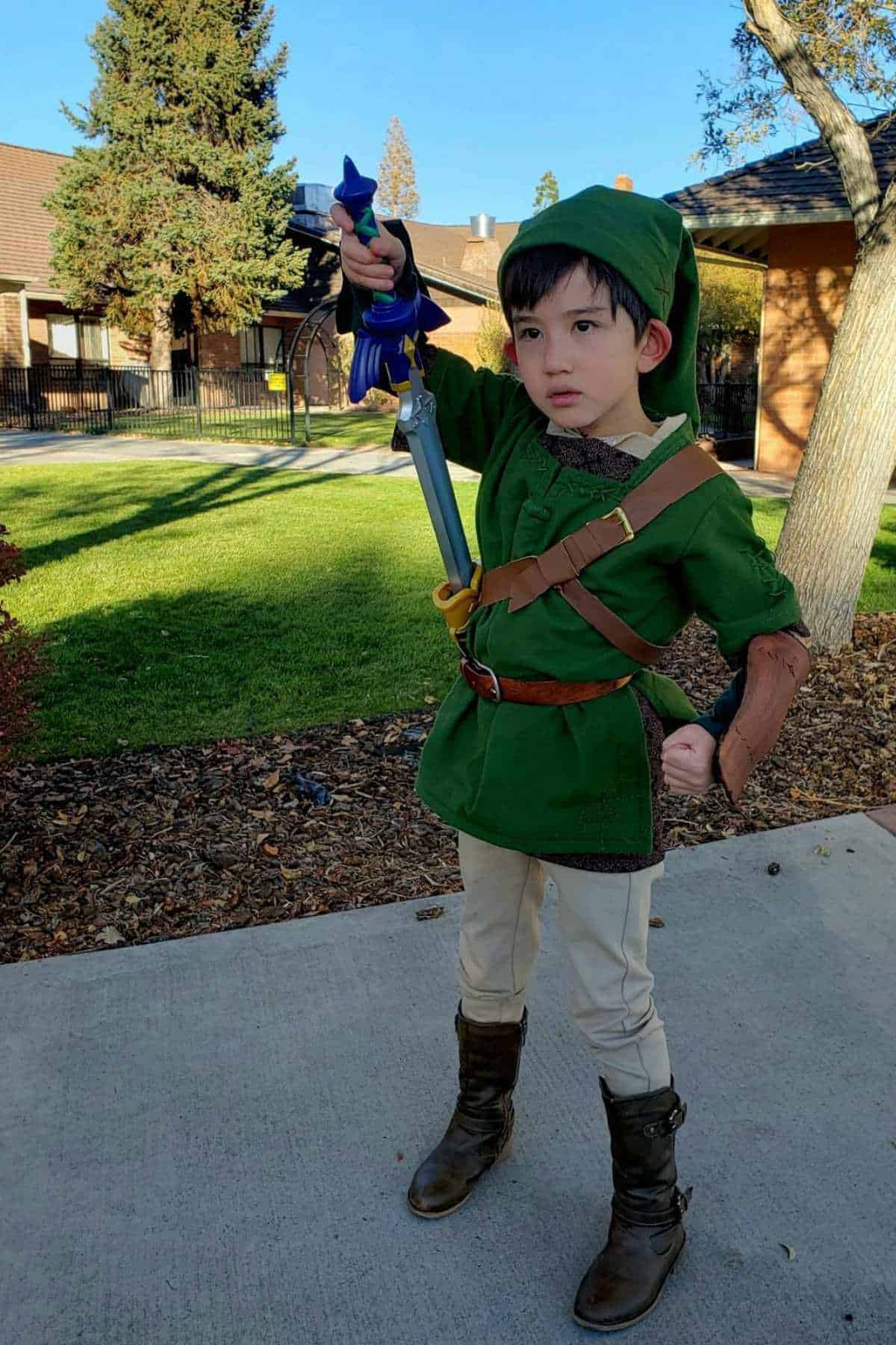 A child dressed as a Link character in a green tunic, hat, and boots holds a toy sword outside on a sidewalk with grass and trees in the background.