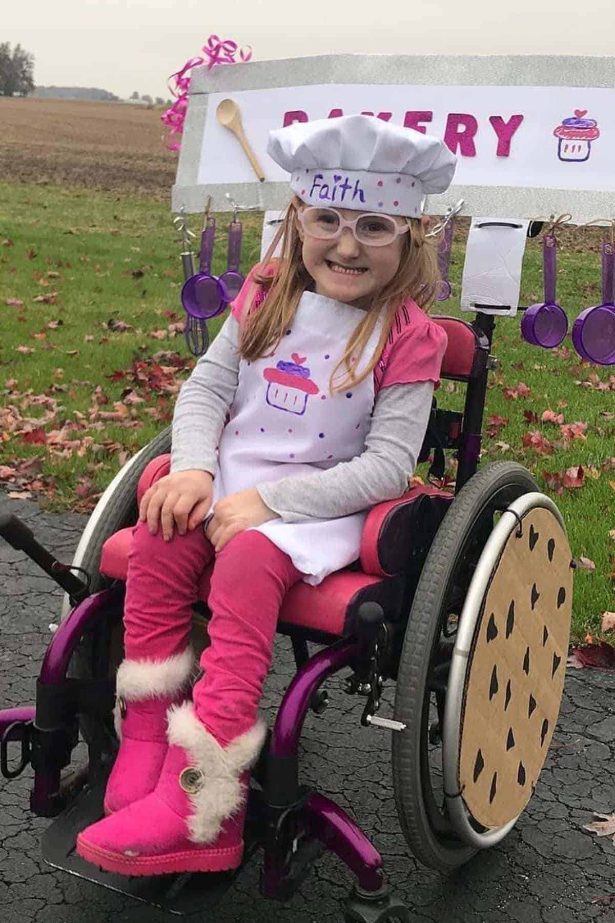 A young girl in a wheelchair, dressed as a baker with a hat and apron labeled “Faith,” sits in front of a toy bakery stand outdoors.