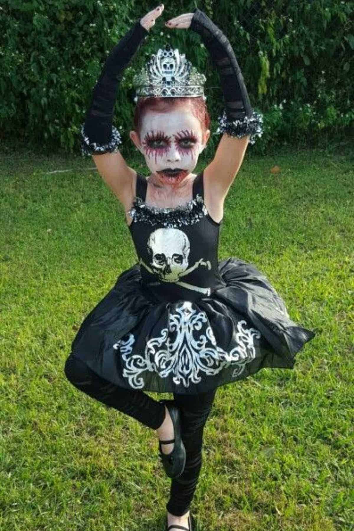 Child in a zombie ballet costume and tiara, with dramatic face paint, poses in a ballet stance on grass with greenery in the background.