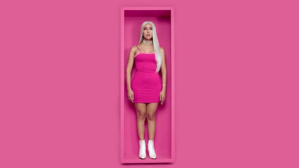 A woman with long blonde hair in a pink dress and white boots stands in a pink, rectangular box against a matching pink background.