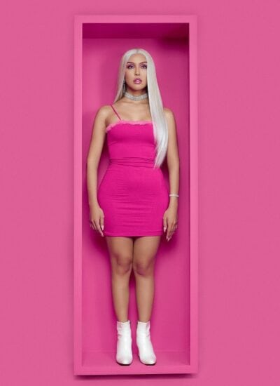 A woman with long blonde hair in a pink dress and white boots stands in a pink, rectangular box against a matching pink background.