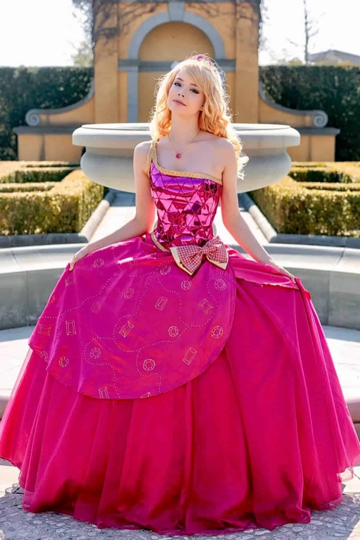 A person wearing Barbie Princess Charm a shiny pink ballgown with a geometric bodice and large bow stands outdoors in front of a fountain and trimmed hedges.