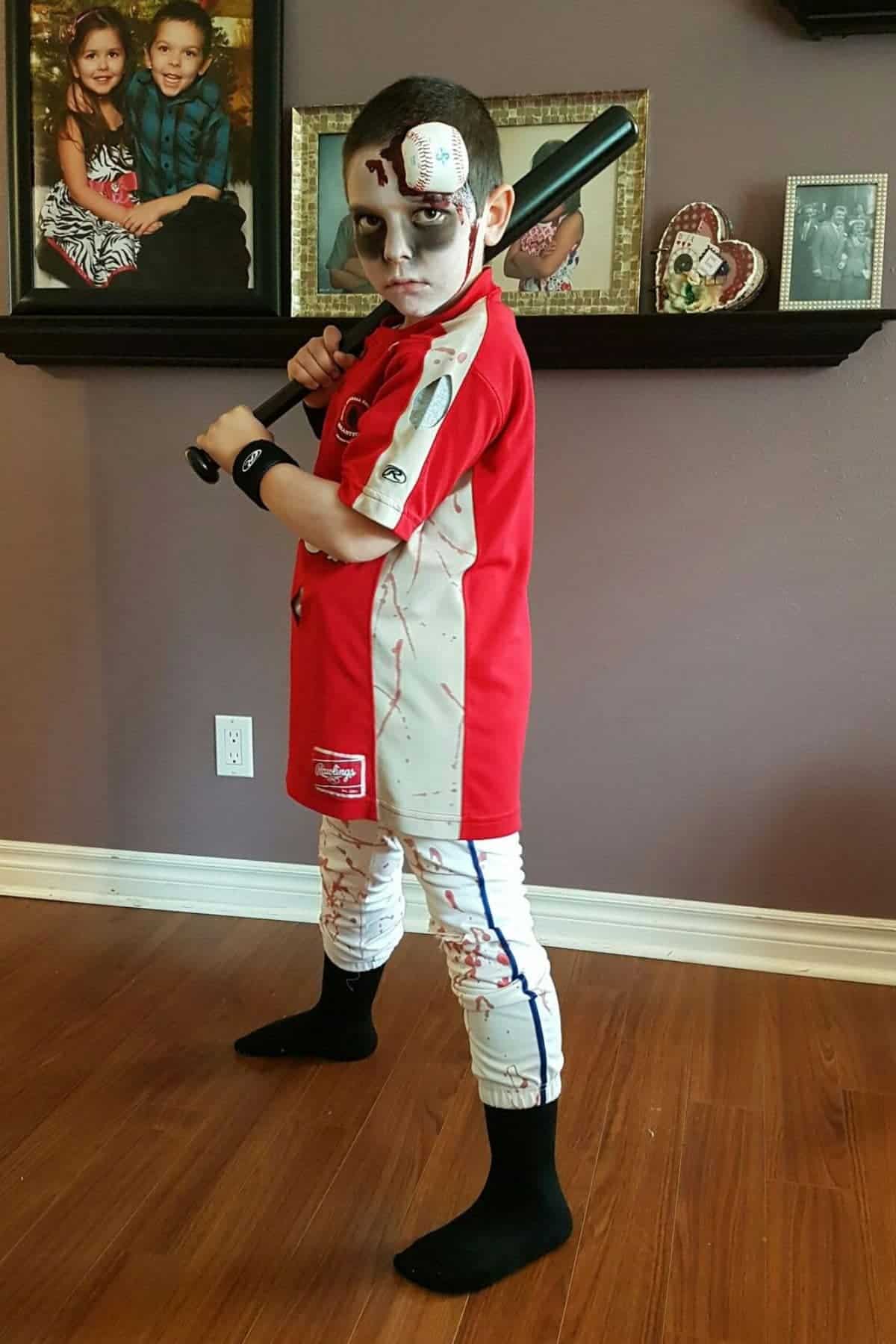 Child in a blood-stained baseball uniform and zombie makeup holds a bat indoors, standing on a wooden floor with family photos on the wall behind.