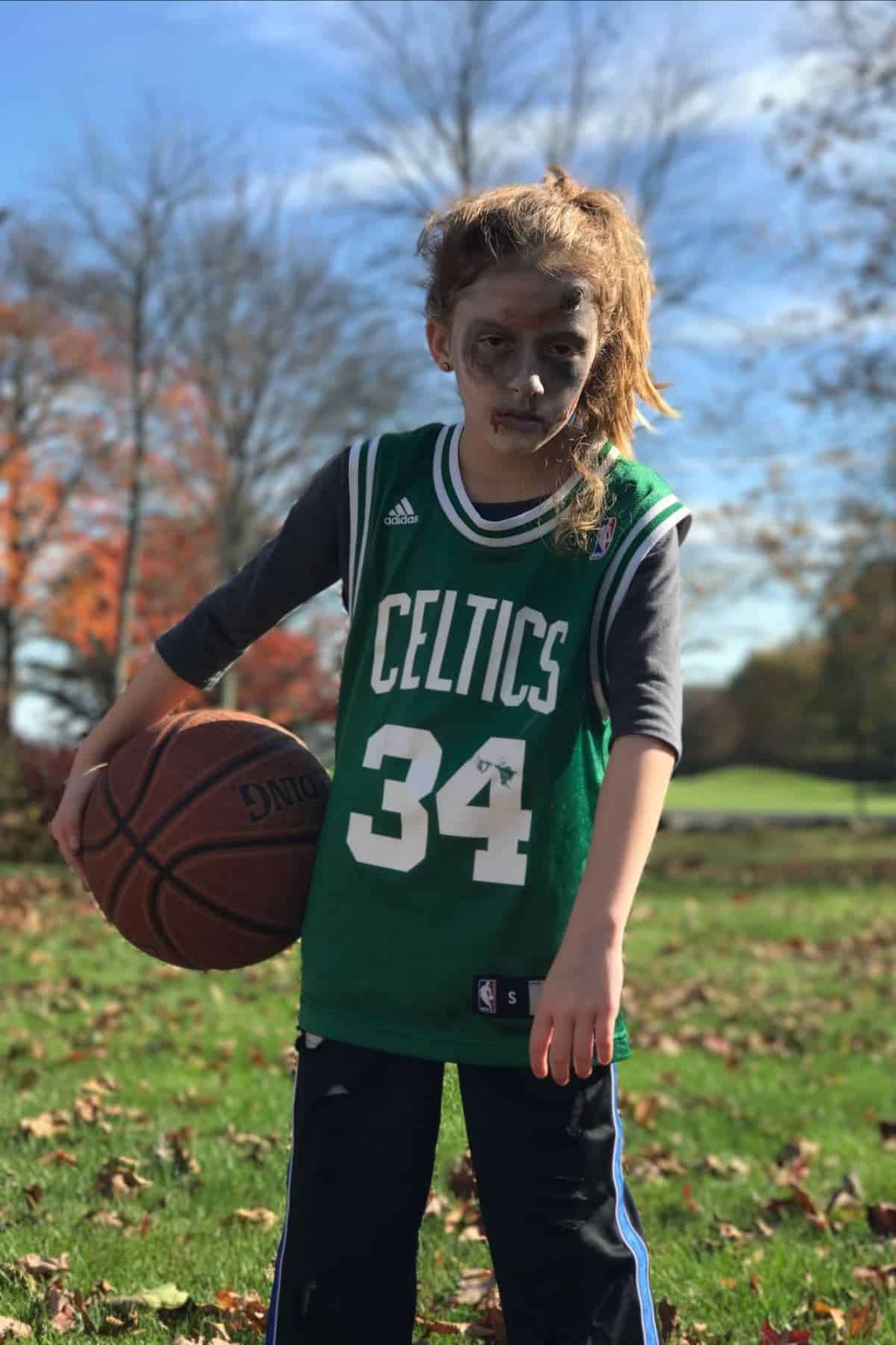 A child in a Celtics jersey with “34” holds a basketball outdoors, wearing face paint that resembles zombie makeup. Autumn trees and blue sky are visible in the background.