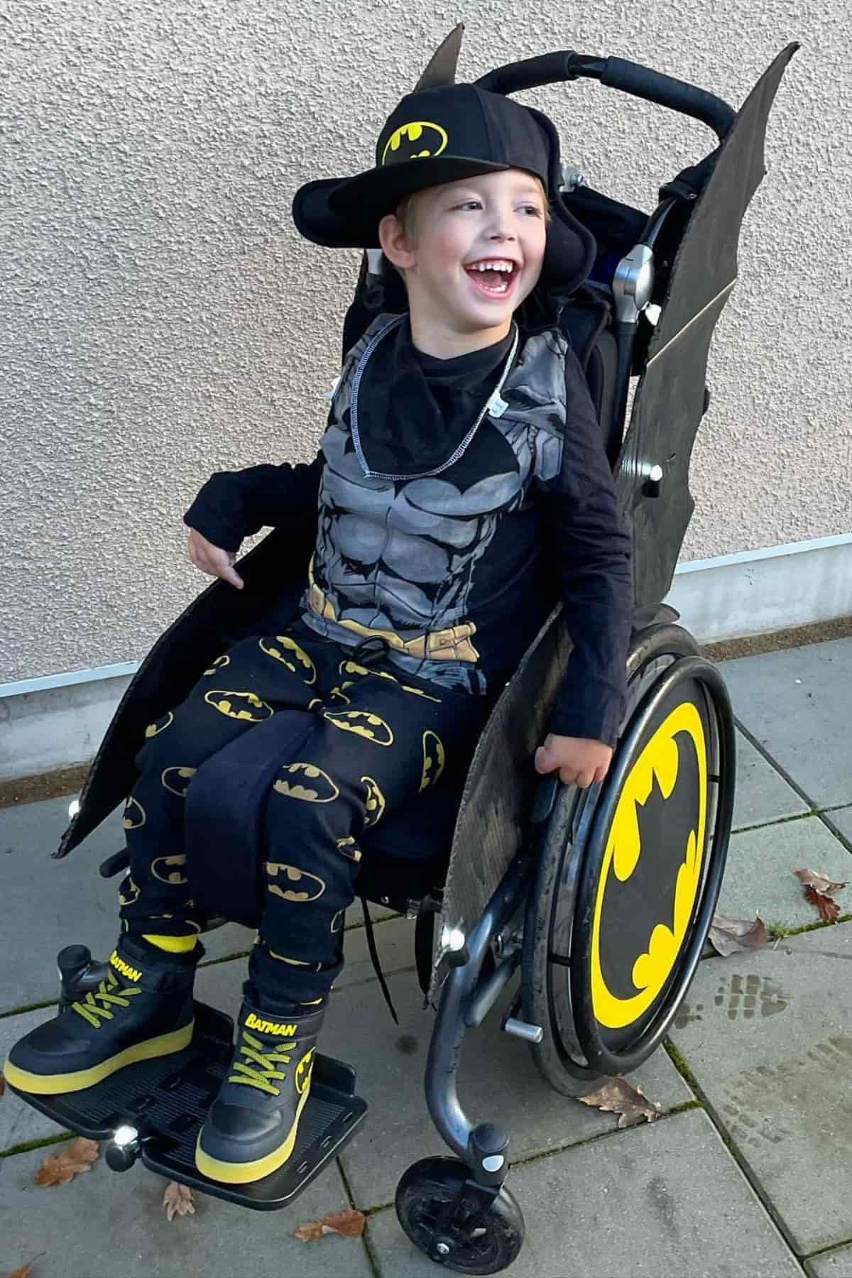 Smiling child in a Batman costume sits in a wheelchair decorated with Batman symbols, outdoors on a paved area.