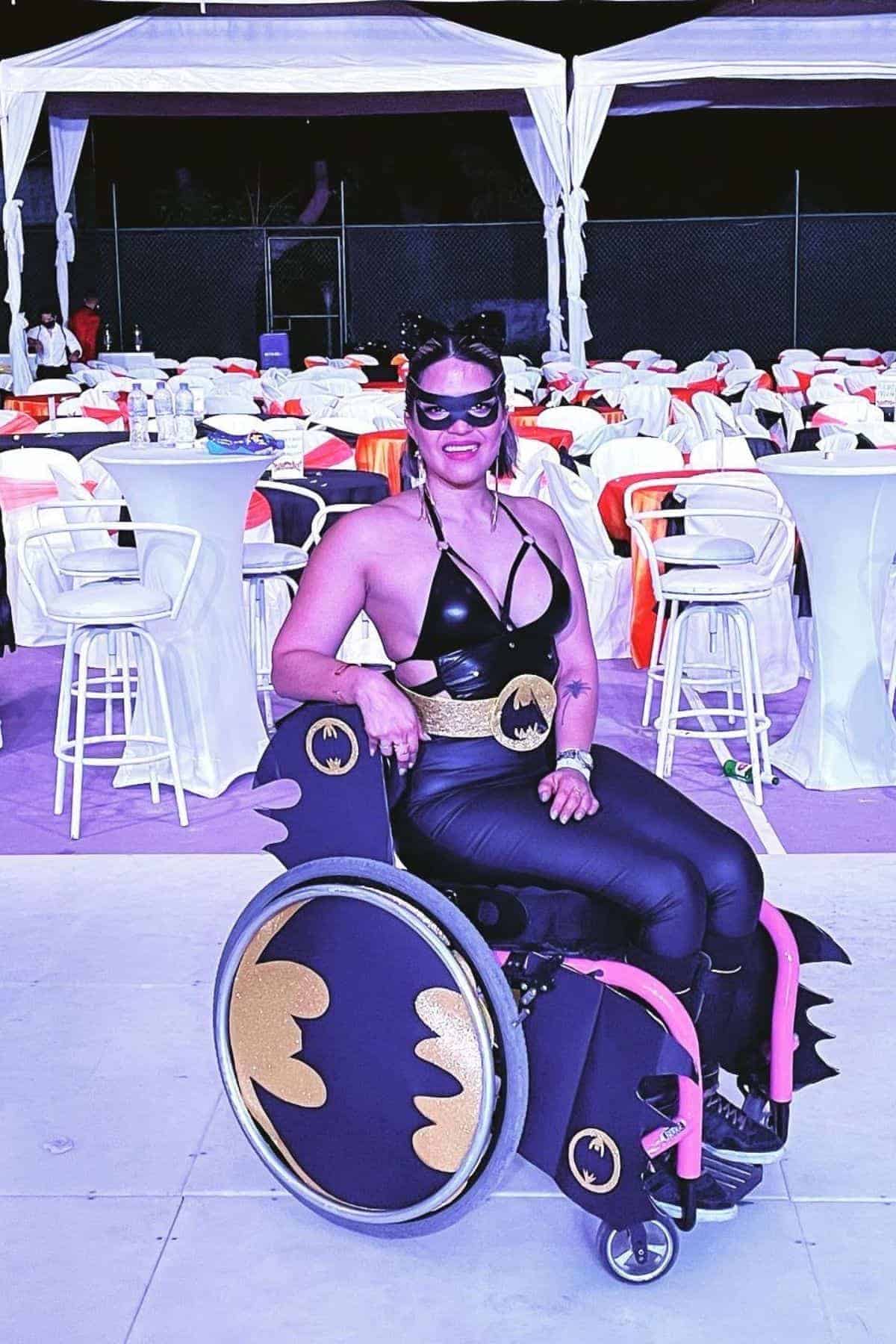 A person in a Batwoman costume sits in a wheelchair decorated with black and gold bat symbols, surrounded by tables and chairs at an event.