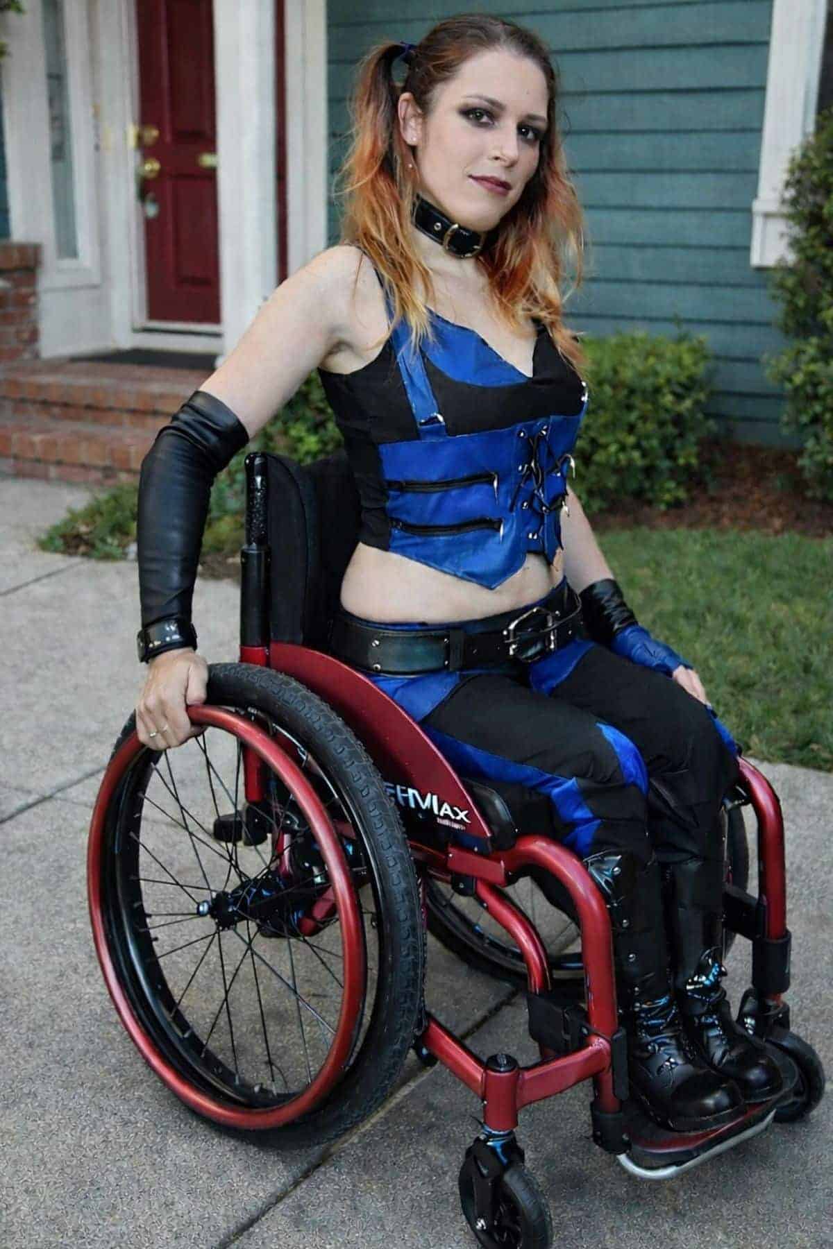 A woman in a red wheelchair wears a blue-black costume with arm bands and boots, posing outdoors on a sidewalk in front of a blue house with a red door.