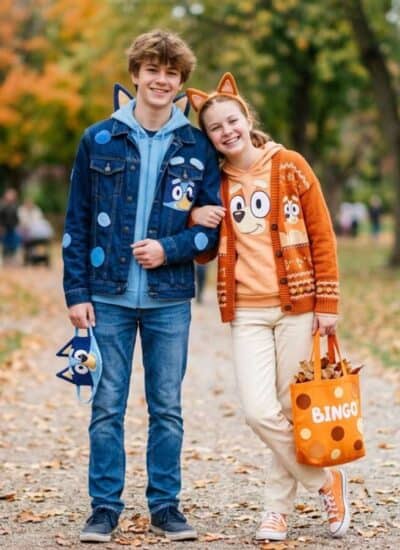 Two people in Bluey and Bingo-themed outfits stand on a leaf-covered park path, smiling, with a group of people and trees with autumn foliage in the background.