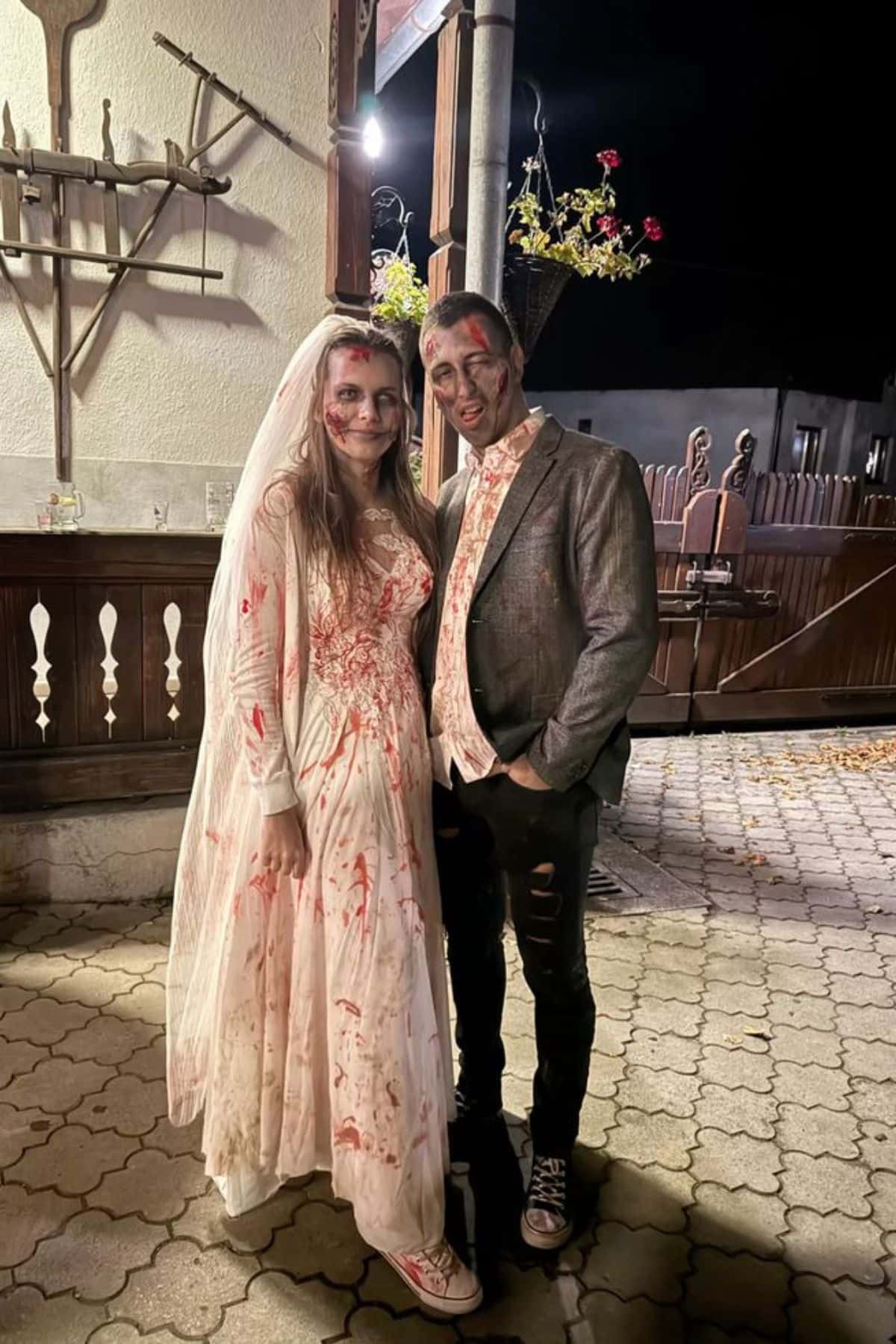 Two people wearing zombie bride and groom costumes with fake blood stand together outdoors at night, smiling for the camera.
