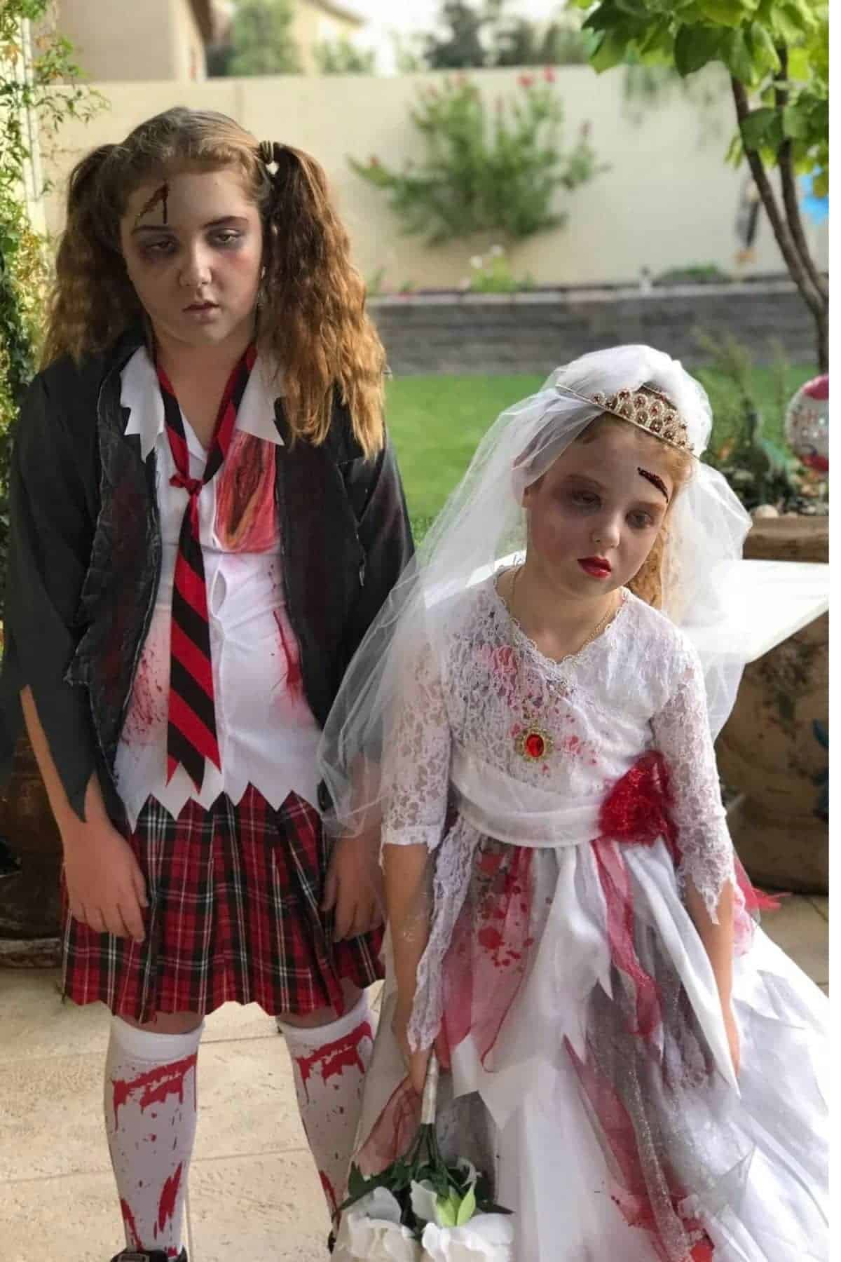 Two children dressed in zombie costumes, one in a tattered school uniform and the other in a blood-stained bridal dress, stand outdoors with serious expressions.