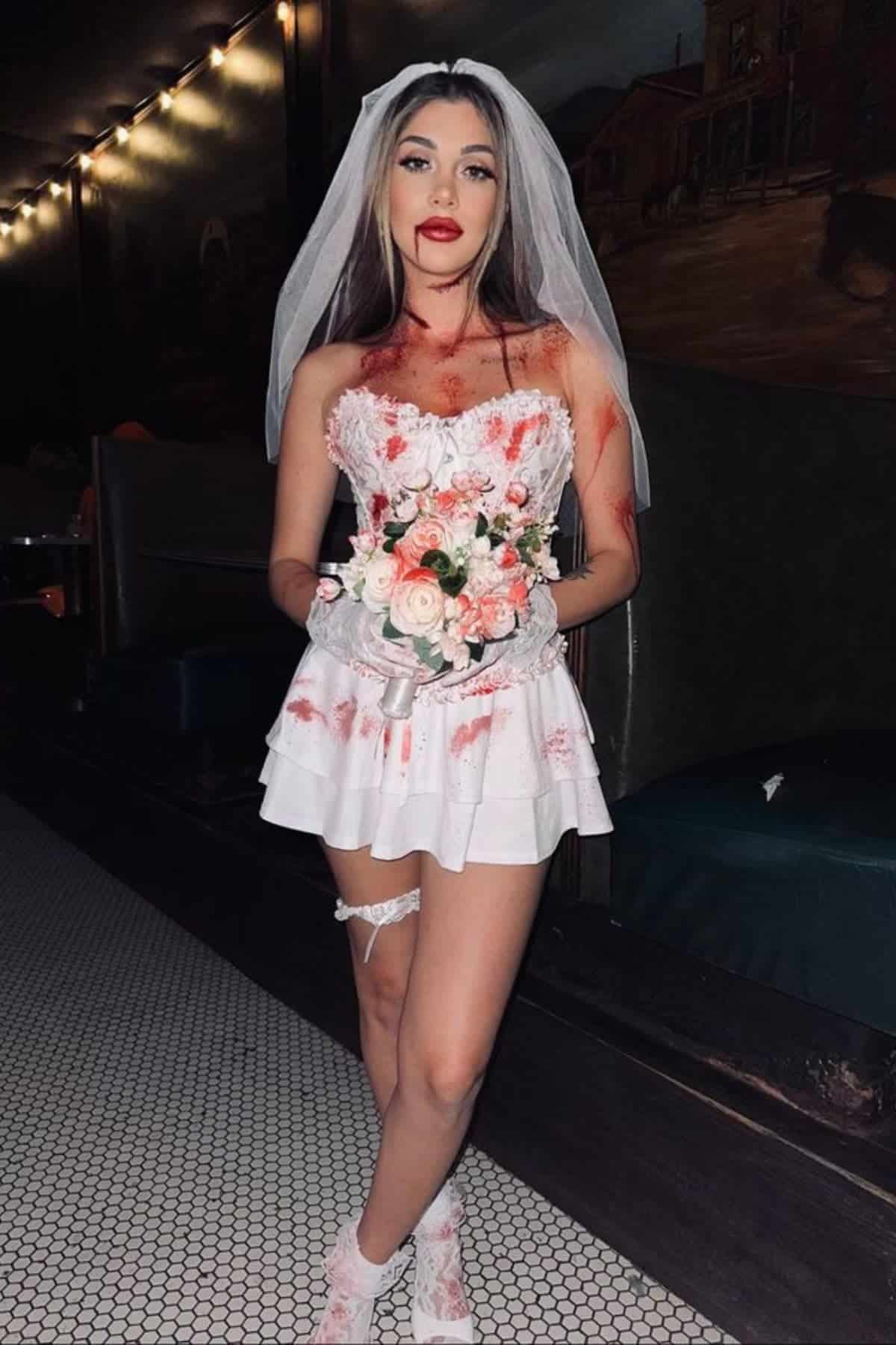 A woman in a short white wedding dress and veil, holding a bouquet, stands indoors with fake blood on her outfit and skin.