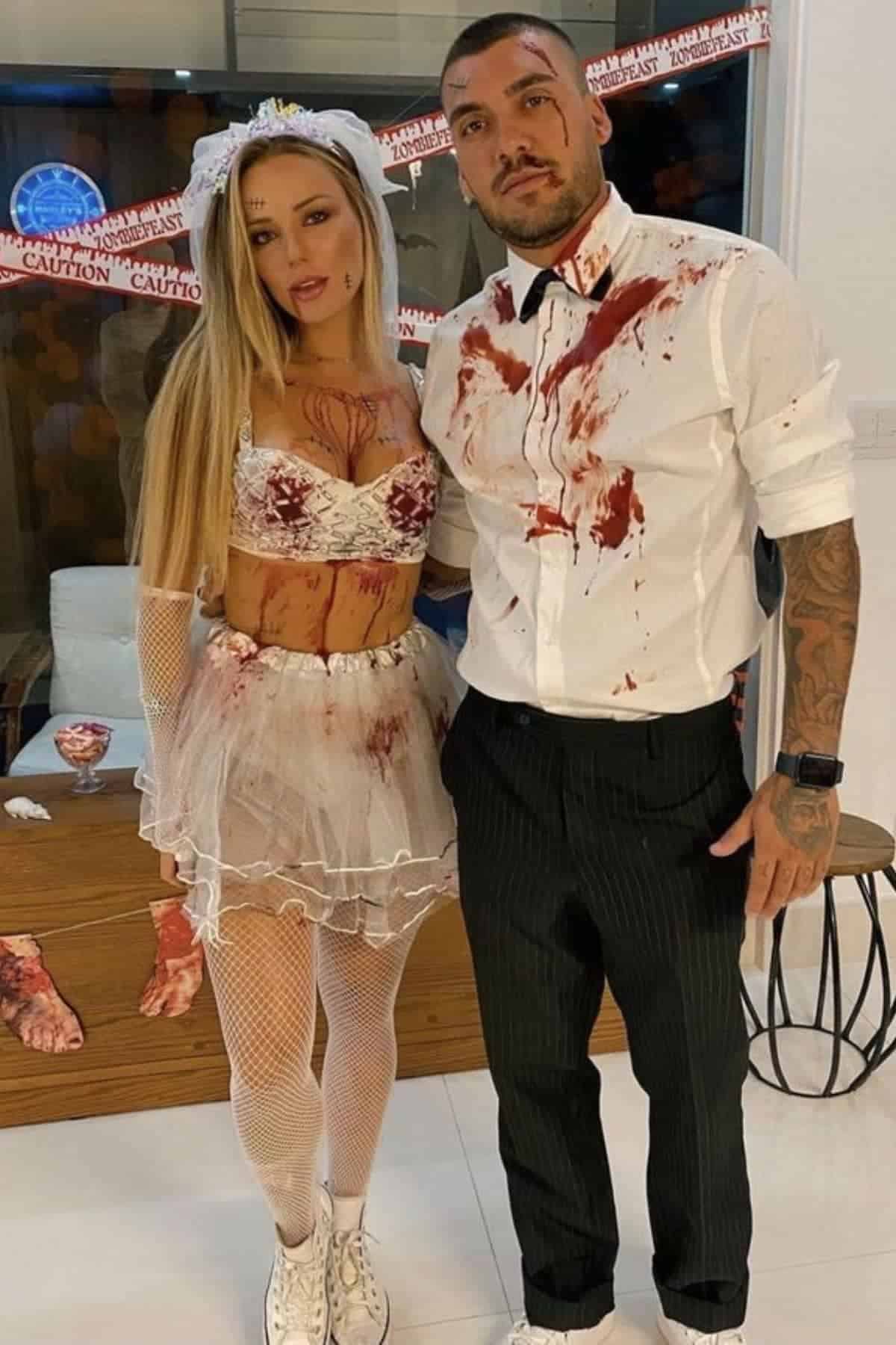 Two adults in Halloween costumes with fake blood; one in a white dress with a veil, the other in a white shirt and black pants. “Caution” tape and props are visible in the background.