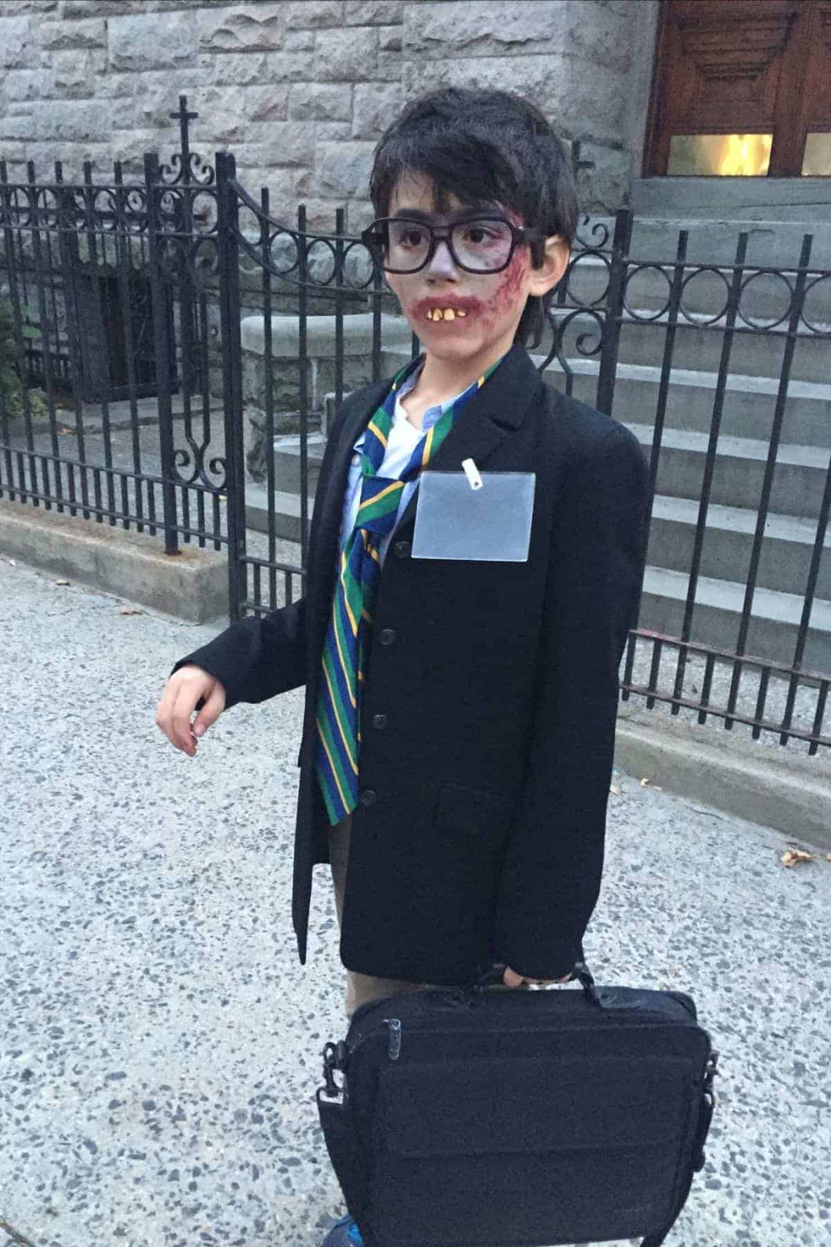 Child in a suit with a striped tie, name tag, briefcase, large fake glasses, and fake teeth, standing on a sidewalk in front of stone steps and a black iron fence.