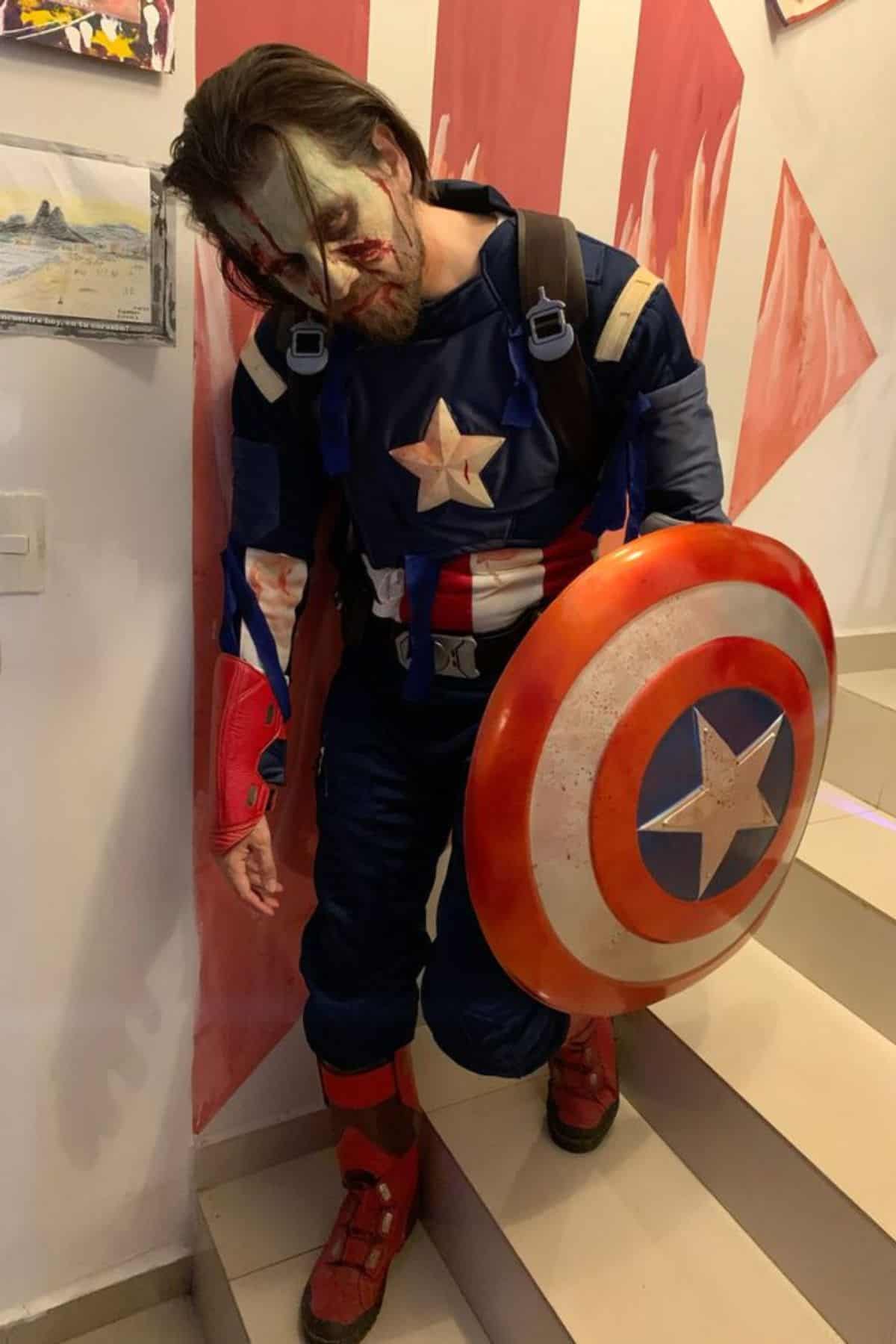 Person in a worn Captain America costume with zombie-like makeup stands on stairs, holding a shield and wearing a backpack.