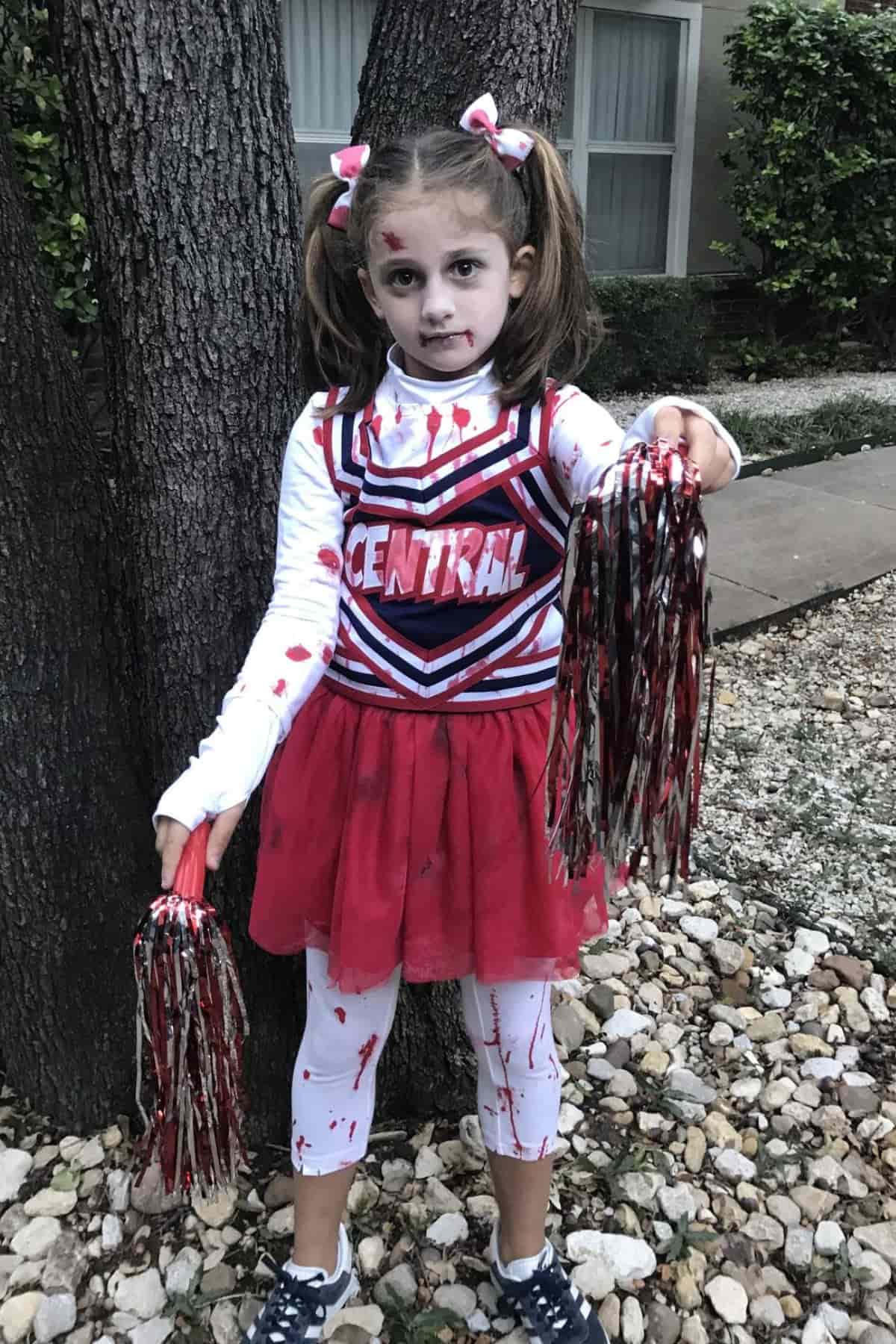 Child in a blood-splattered cheerleader costume, holding red and black pom-poms, standing outdoors near a tree and rocks.