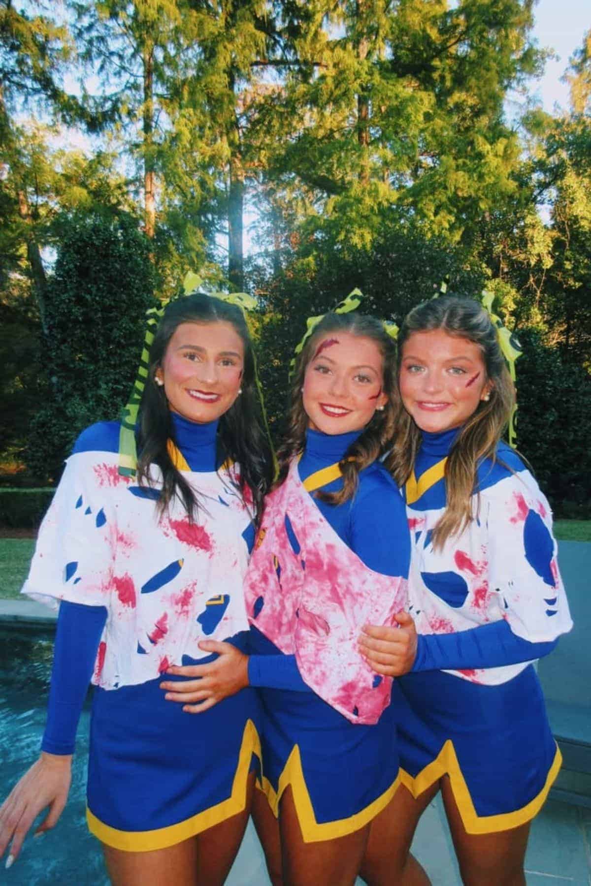 Three people in blue and yellow cheerleader uniforms with fake blood stains stand outdoors, smiling with arms around each other, against a background of green trees.