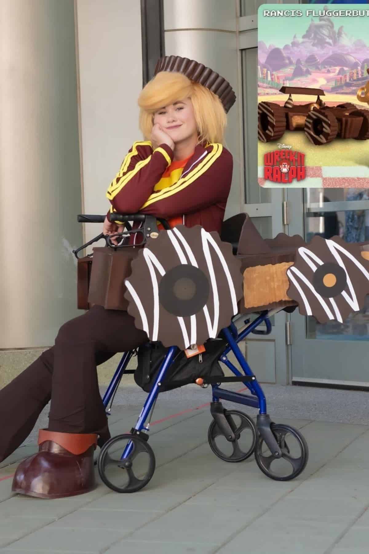 Person in a wheelchair cosplays as a character with cookie and chocolate-themed props, wearing a yellow wig and brown costume, seated outdoors near a Wreck-It Ralph poster.