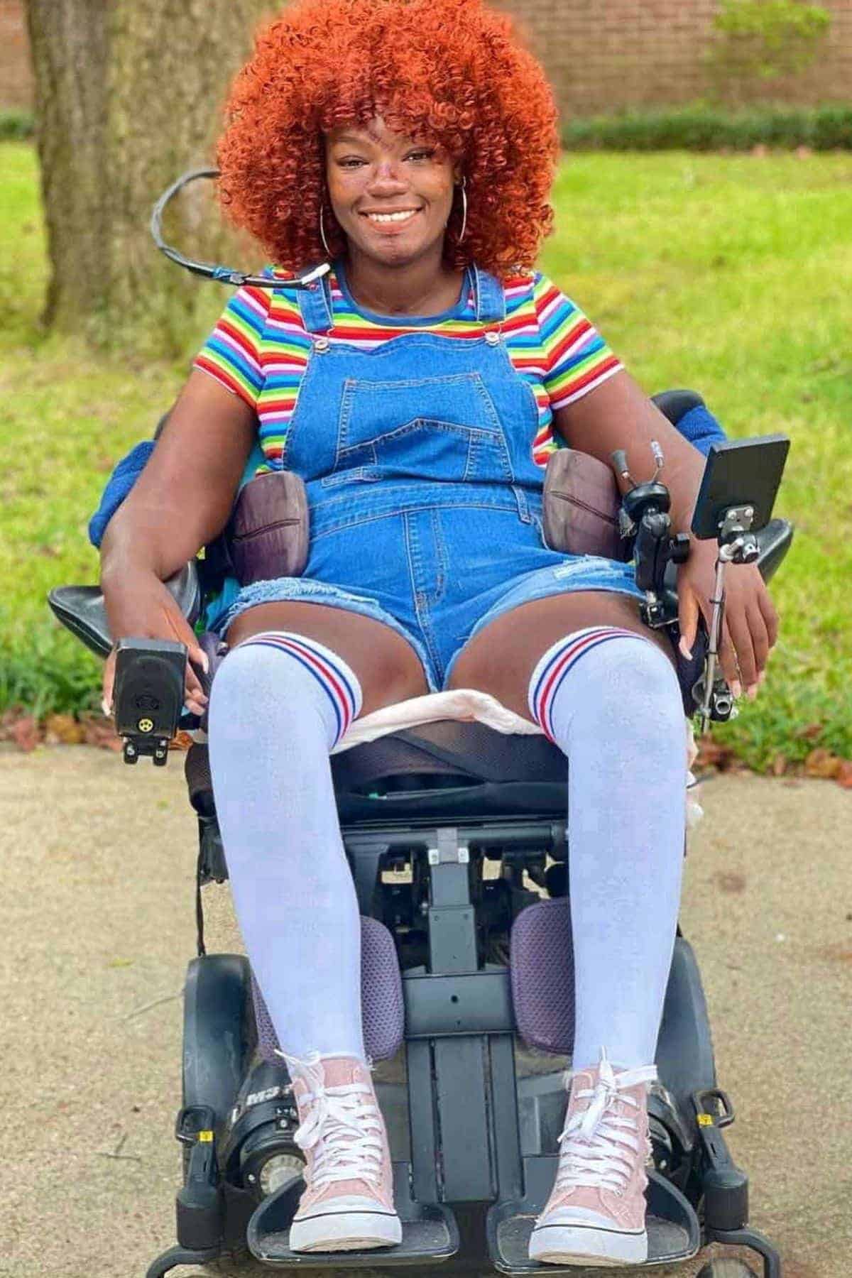 A woman with red curly hair, wearing a striped shirt, denim overalls, and white knee-high socks sits in a power wheelchair outdoors on a sidewalk.
