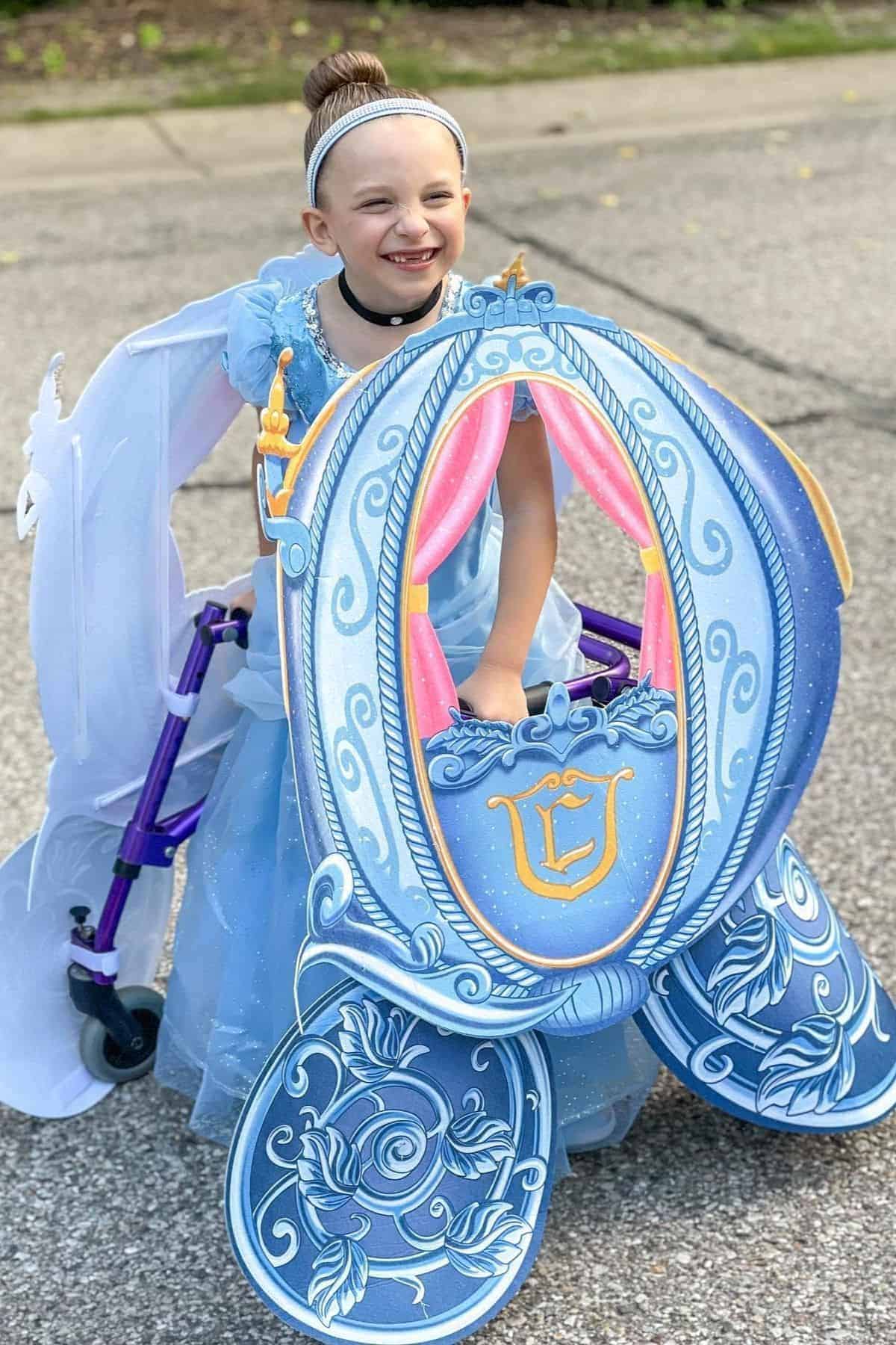 A young girl in a blue princess costume uses a walker decorated to look like a royal carriage, smiling outdoors on a paved surface.