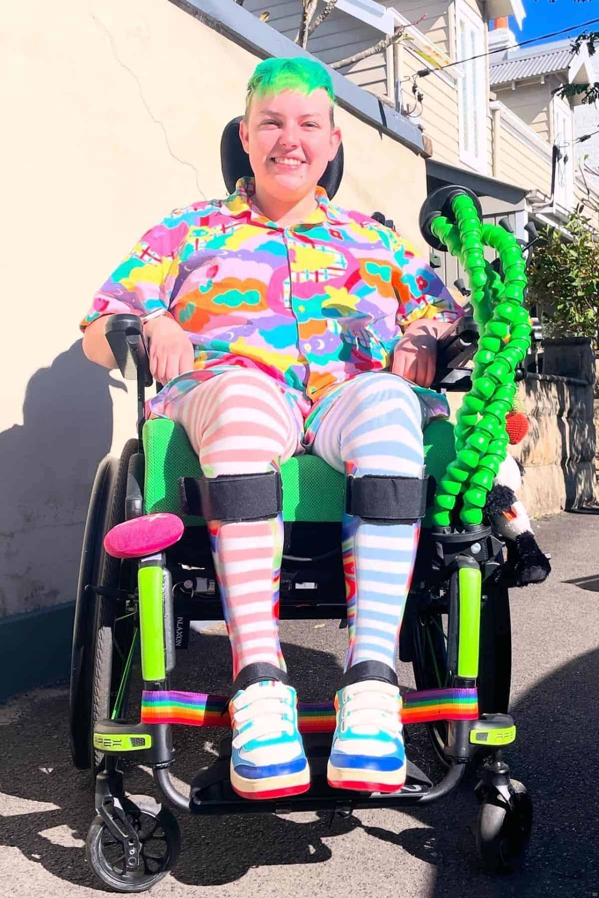 A person with short green hair sits in a wheelchair, wearing colorful patterned clothes and striped leg warmers, smiling outdoors on a sunny day.