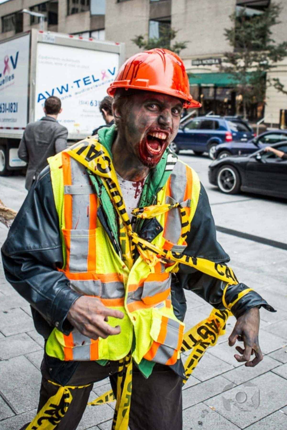 Person in a construction worker outfit with fake blood and zombie makeup, wrapped in caution tape, standing on a city street during daytime.