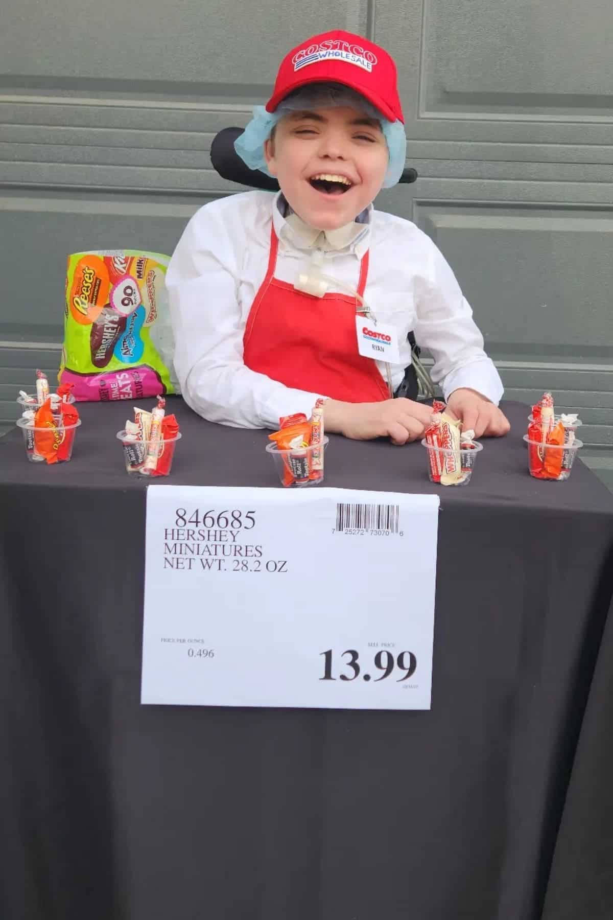 A smiling child in a red cap and apron sits at a table displaying candy samples and a price sign reading “Hershey Miniatures 28.2 oz $13.99.”.
