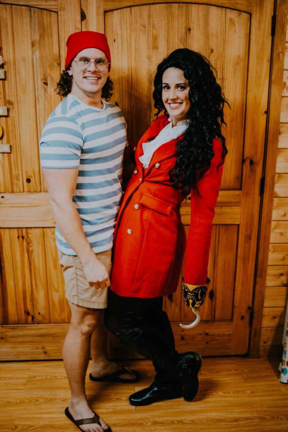 Two people pose indoors; one wears a red beanie, striped shirt, shorts, and flip-flops, the other has a red coat, white scarf, black boots, and a hook hand accessory.
