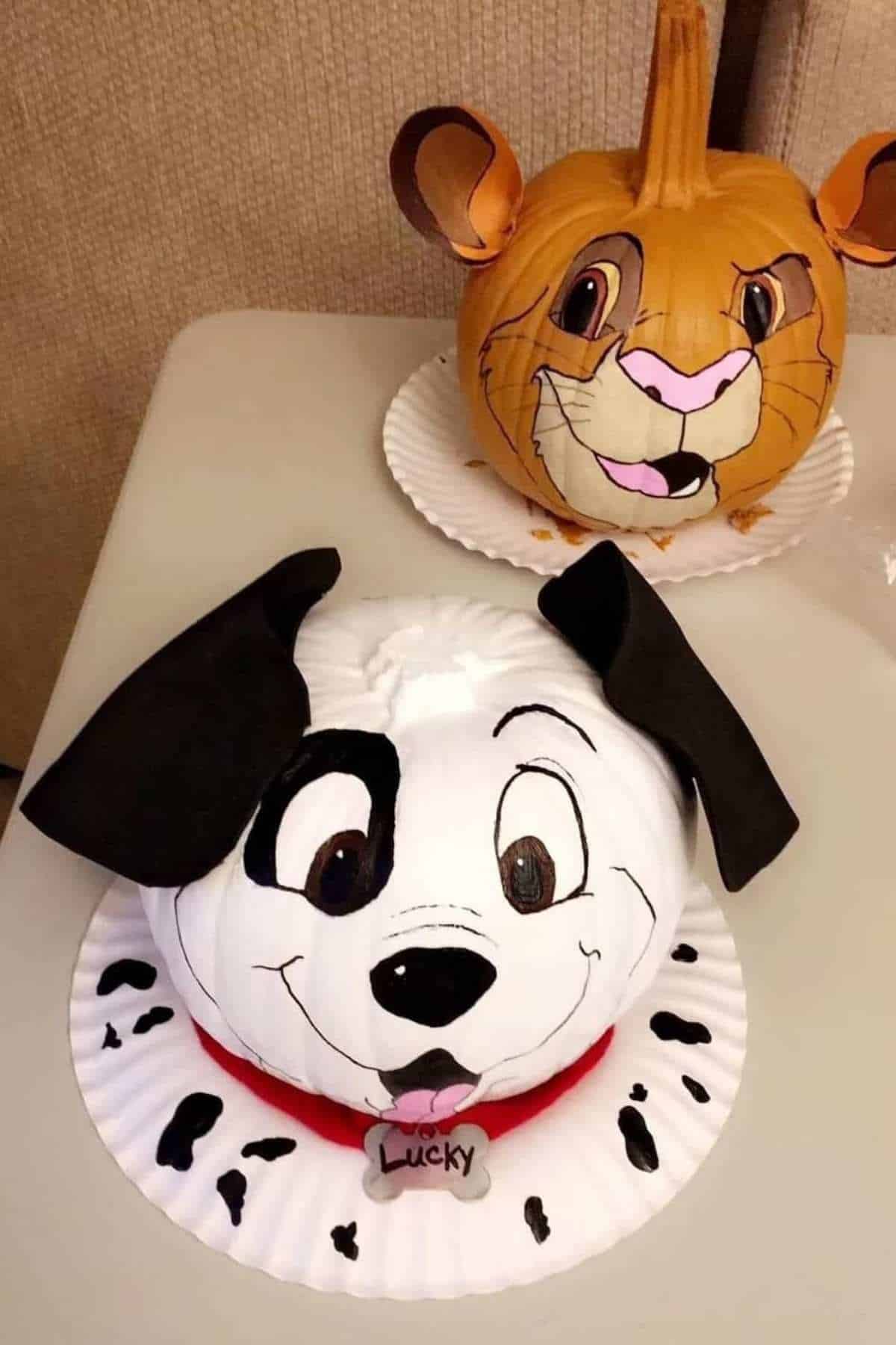 Two pumpkins painted as cartoon animals: a Dalmatian dog with floppy ears in front, and a lion with a smiling face and mane behind, both displayed on white paper plates.