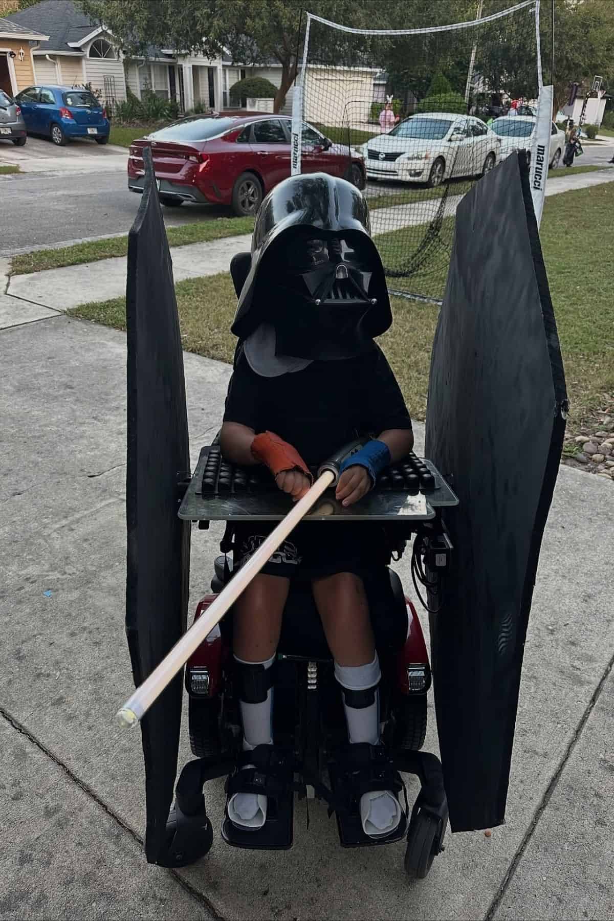Child in a wheelchair dressed as Darth Vader with large black side panels and a lightsaber, outdoors on a residential street.