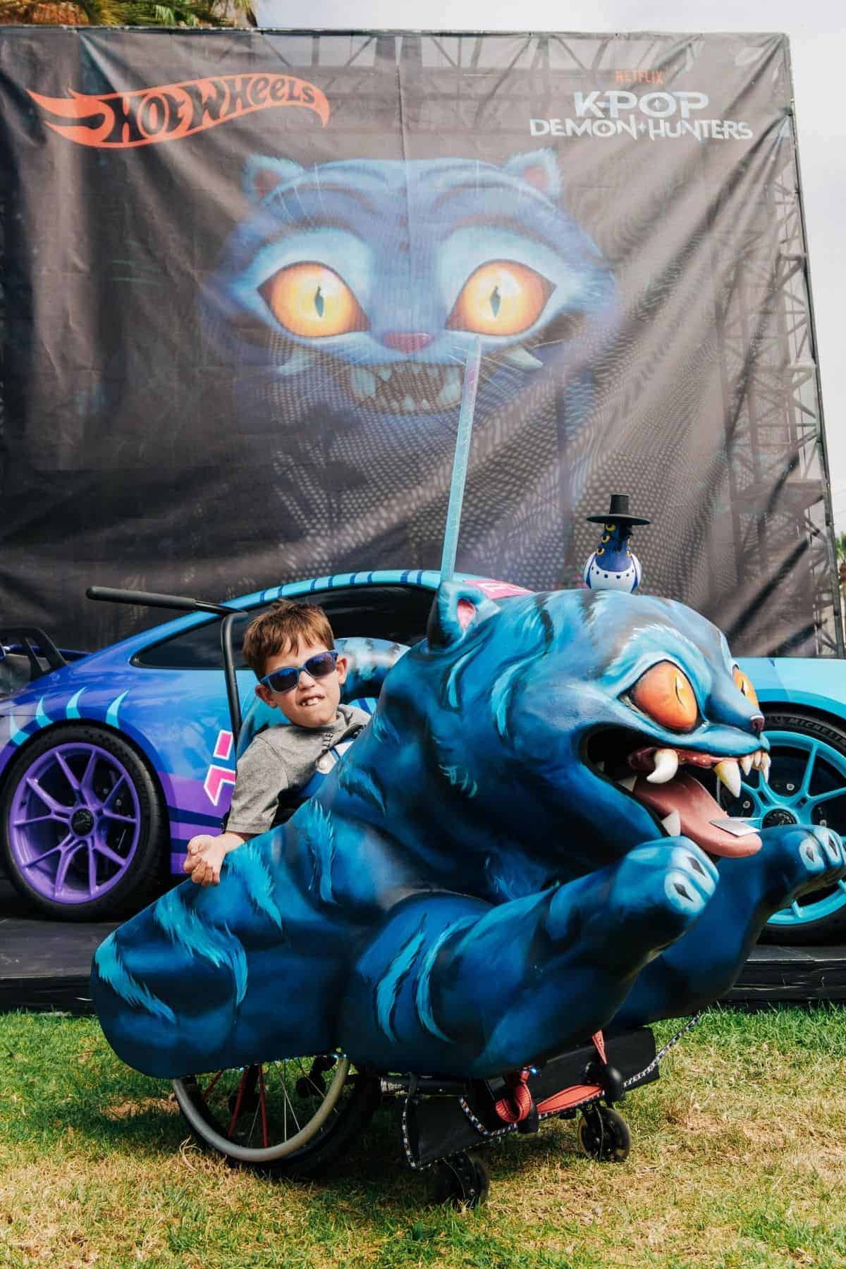 A young boy in sunglasses sits in a wheelchair decorated as a blue monster cat, in front of a matching themed car and large cat backdrop.