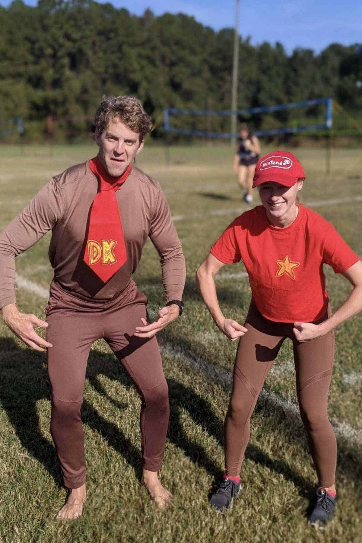 Two people in brown and red costumes pose outdoors on grass, imitating Donkey Kong and Diddy Kong from the Nintendo video game series. A volleyball net is visible in the background.