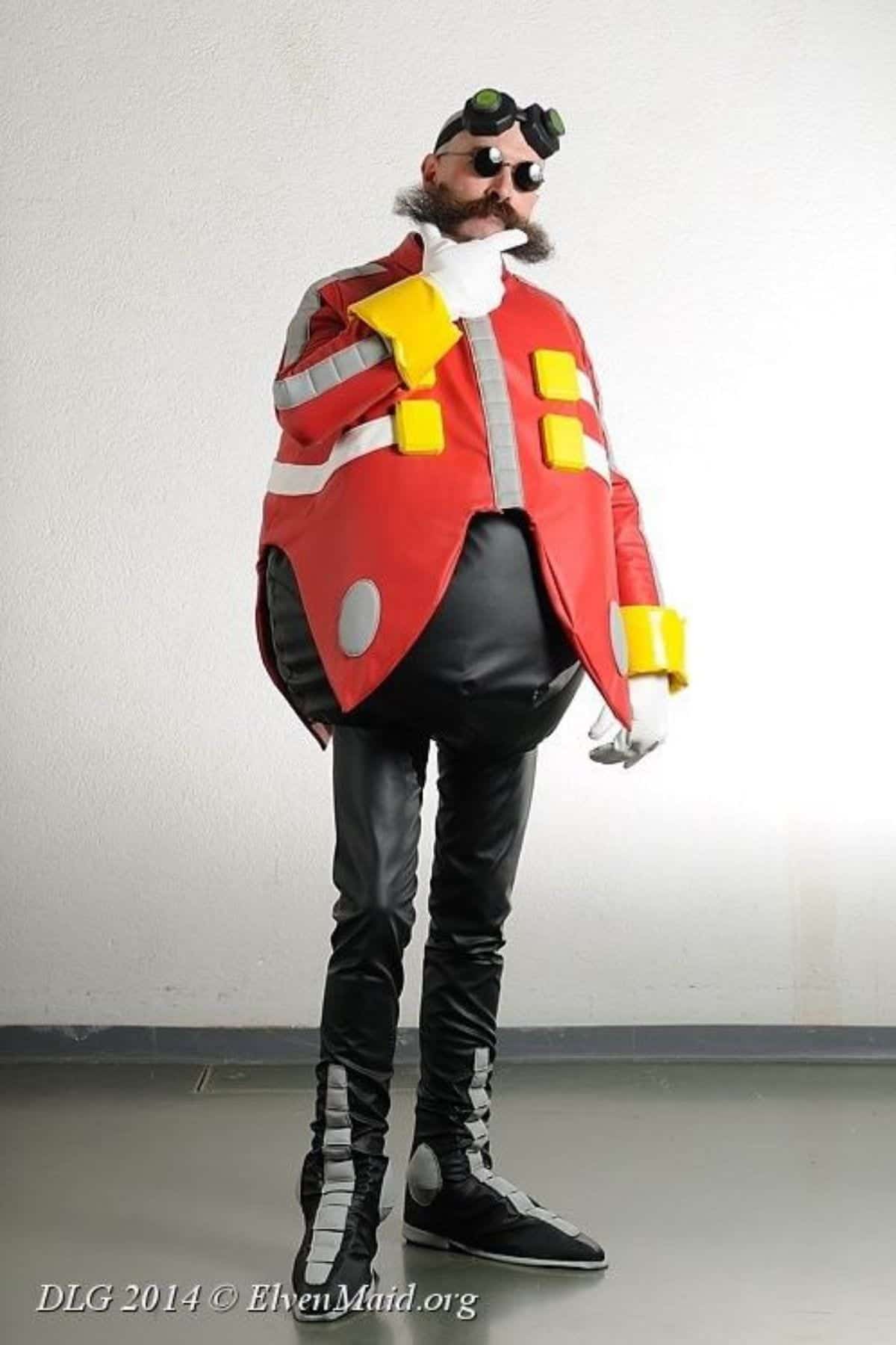 Person in Doctor Eggman costume with a round torso, black pants, boots, gloves, goggles, and a fake mustache, posing indoors against a plain wall.