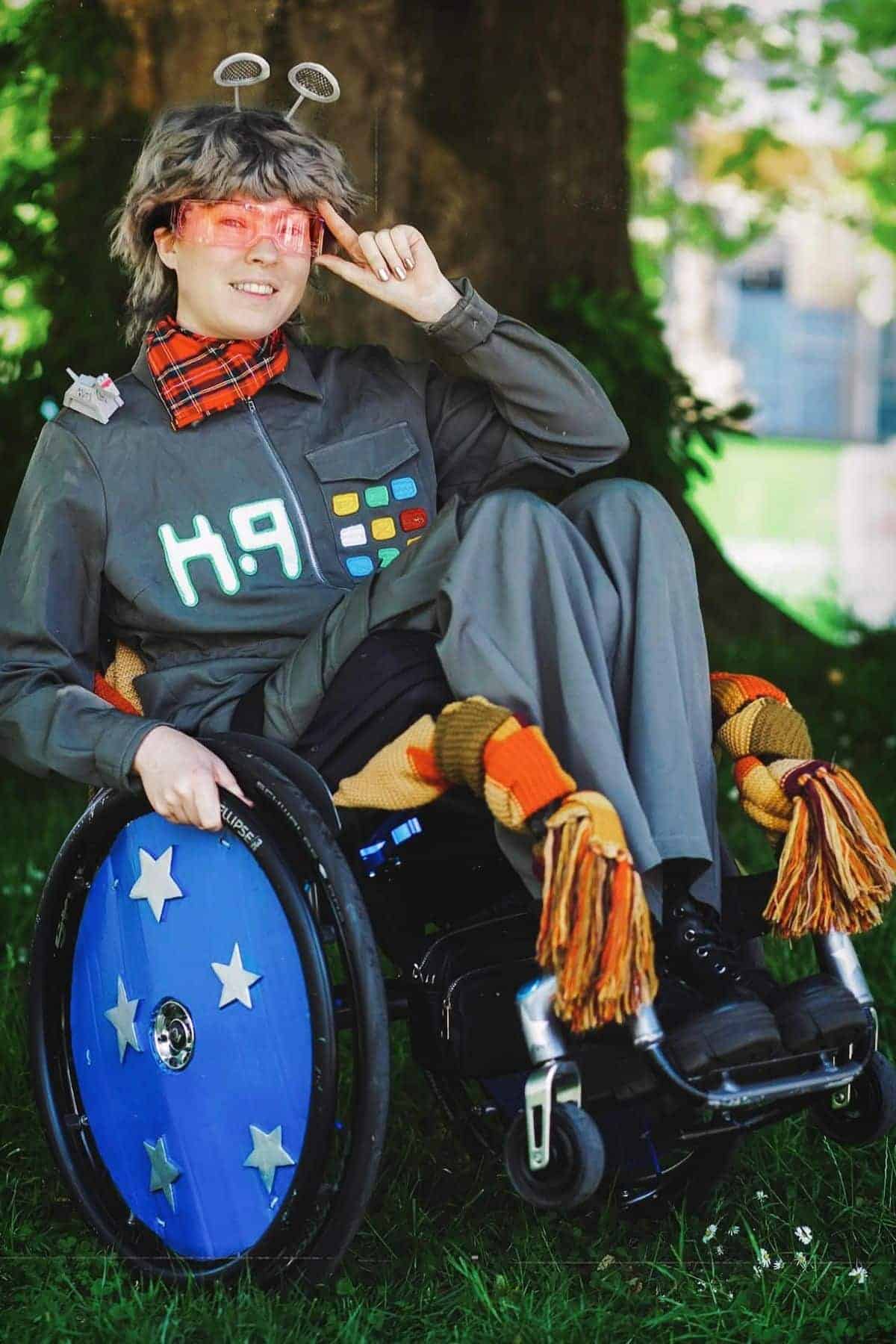 Person in a wheelchair wearing a gray sci-fi-themed outfit, red visor glasses, and a scarf, posing with a peace sign outdoors on grass.