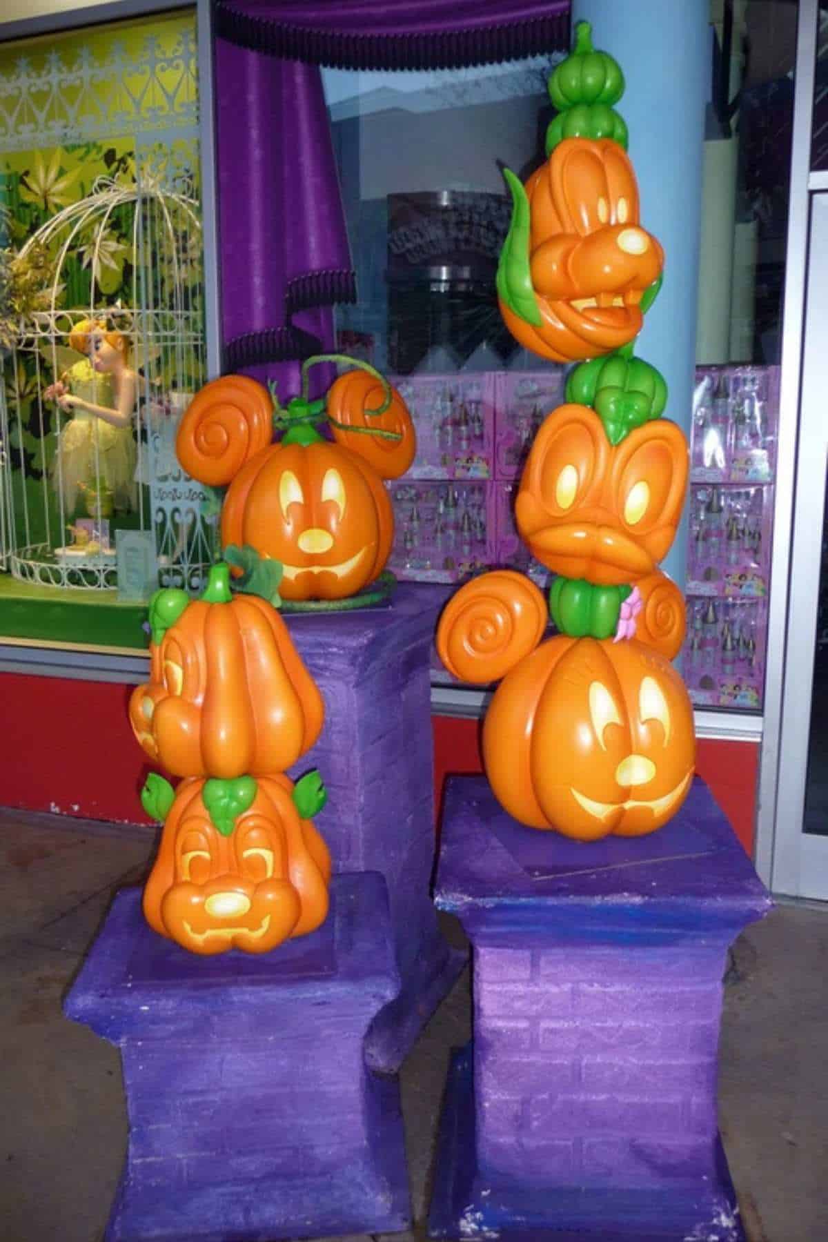 Four pumpkin sculptures carved with cartoon character faces are displayed on purple stands in front of a store window and a decorative wrought iron element.