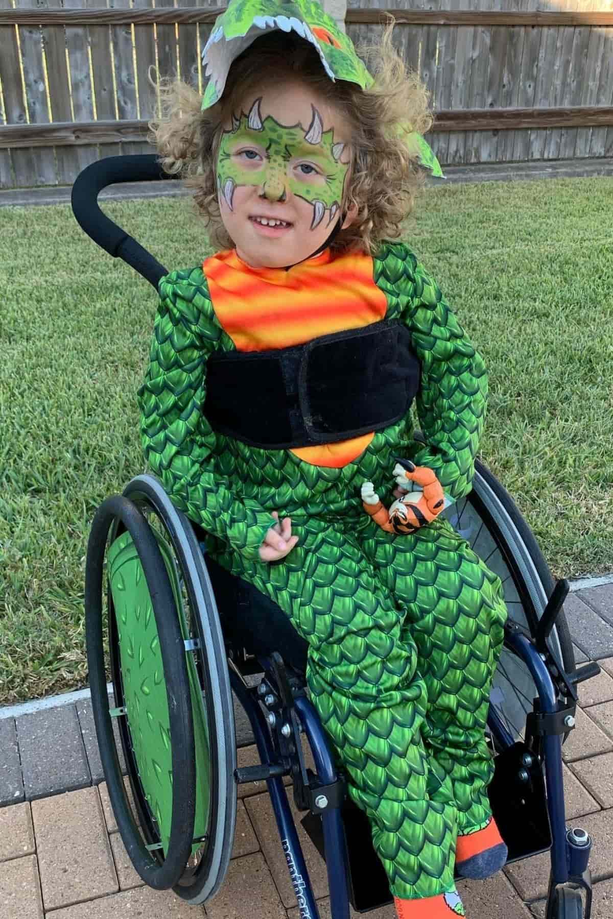 Child in a wheelchair wears a green dinosaur costume and face paint, smiling outdoors on a paved path near grass and a wooden fence.