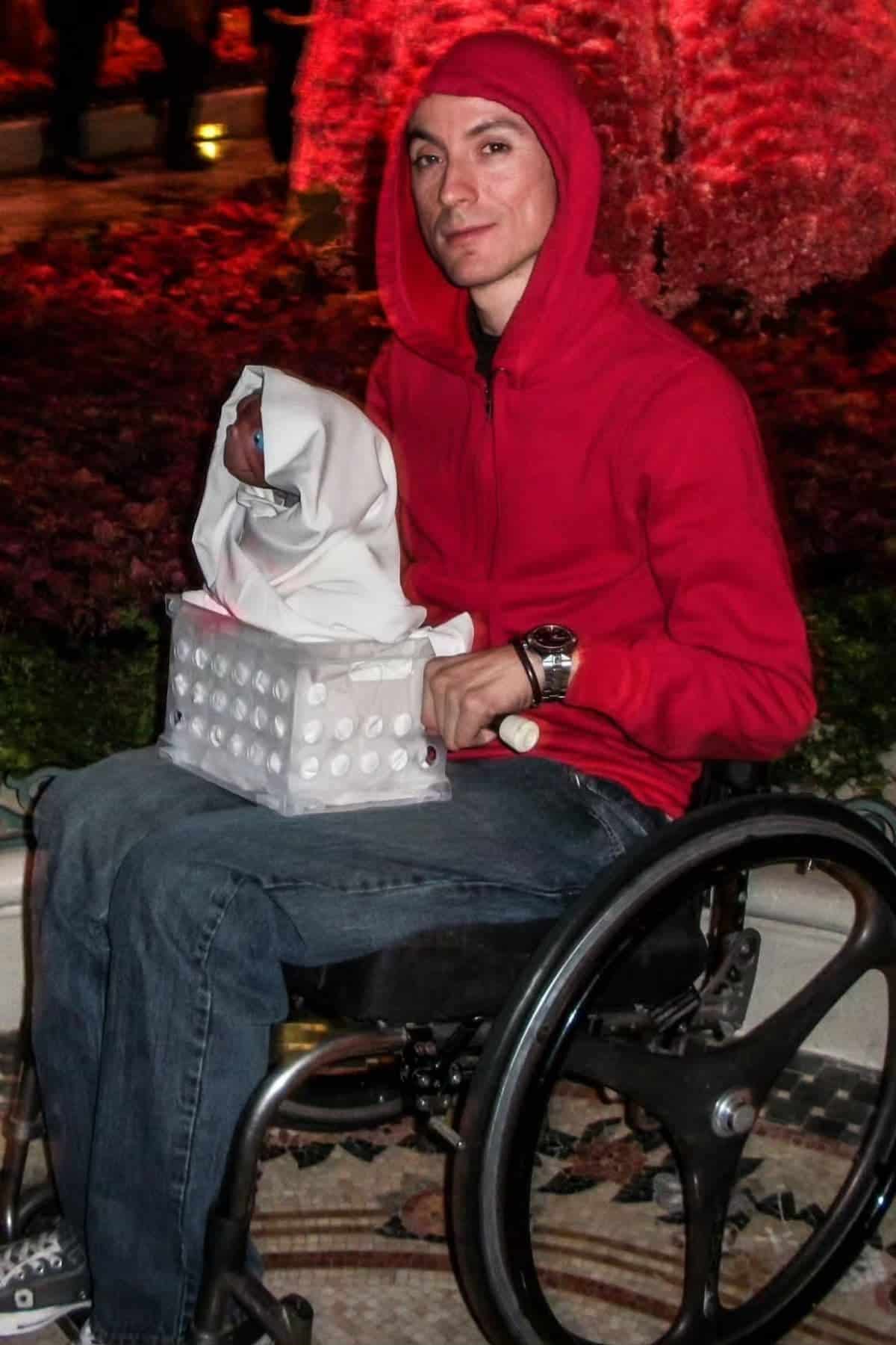 A person in a wheelchair wears a red hoodie and holds a box with a small figure dressed in white, resembling a scene from the movie E.T., outdoors at night.