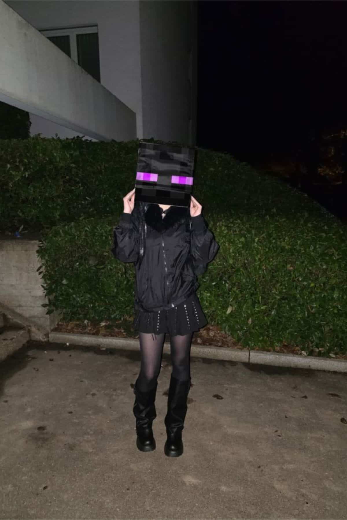 Person wearing a black outfit and black boots stands outdoors at night, holding a box with purple eyes resembling a Minecraft Enderman on their head.
