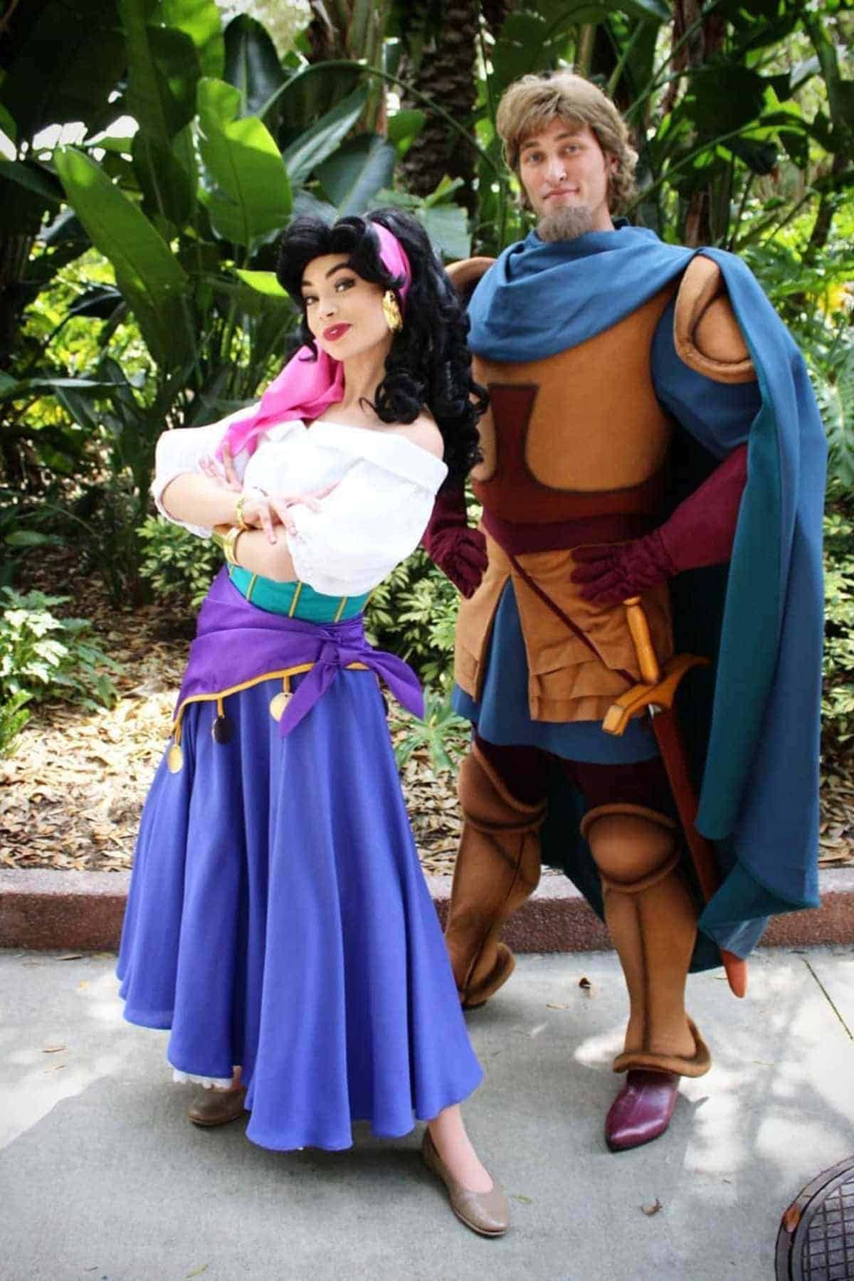 Two people in colorful, medieval-inspired costumes pose outdoors; one wears a white blouse and blue skirt, the other has armor and a cape. Lush green plants are in the background.