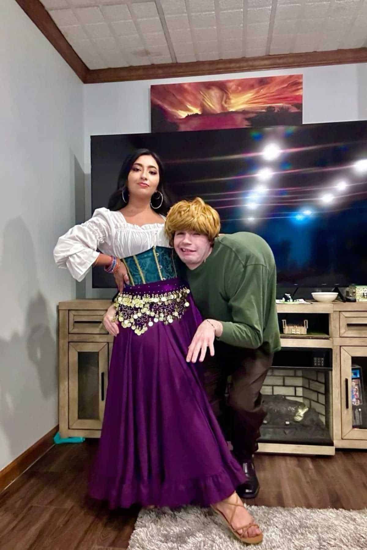 Two people in costume pose indoors; one wears a white blouse, corset, and purple skirt, while the other is dressed in a green top and a red wig, crouching with an arm bent.
