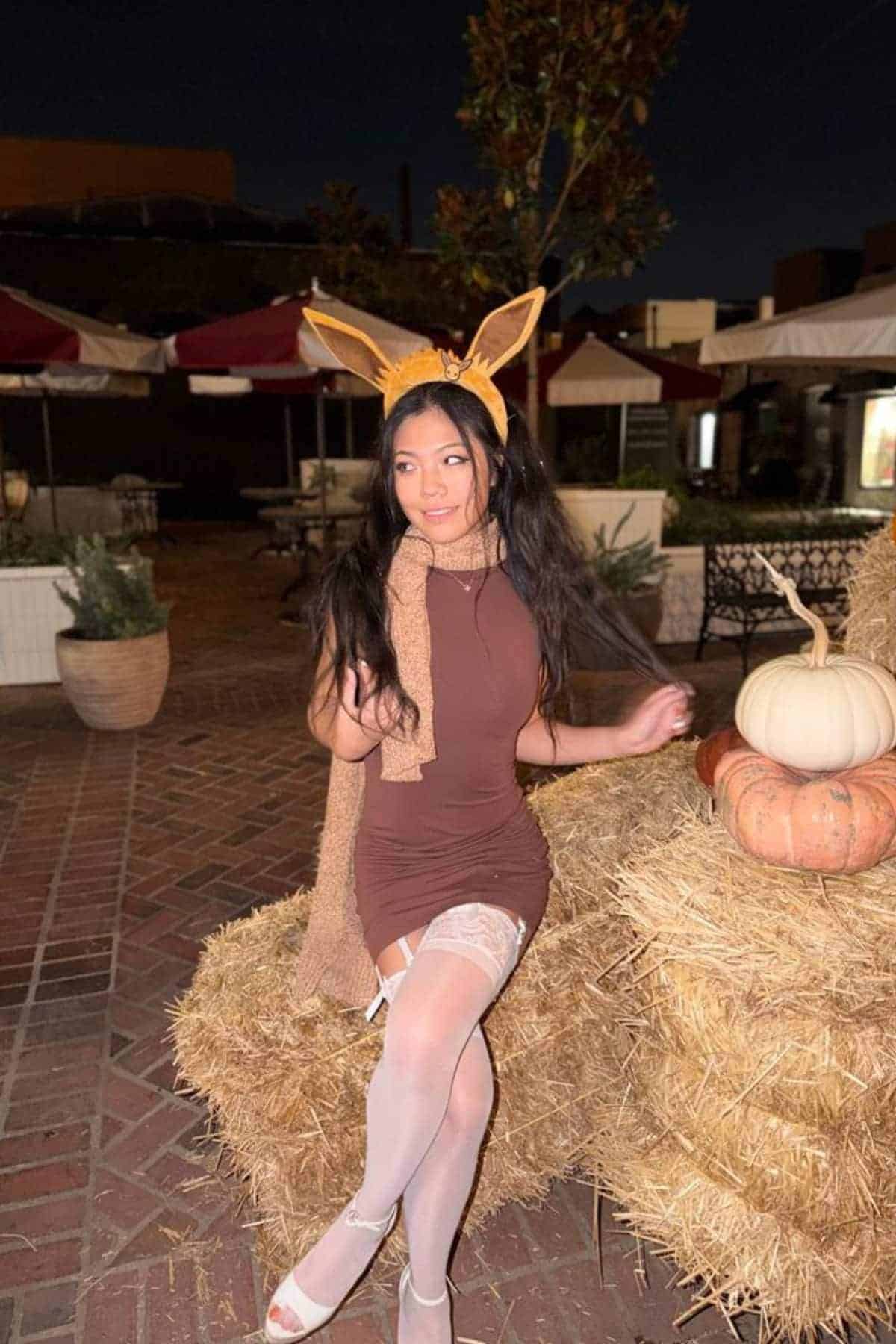 Woman in a brown dress and animal ear headband sits on hay bales next to pumpkins in an outdoor setting at night.