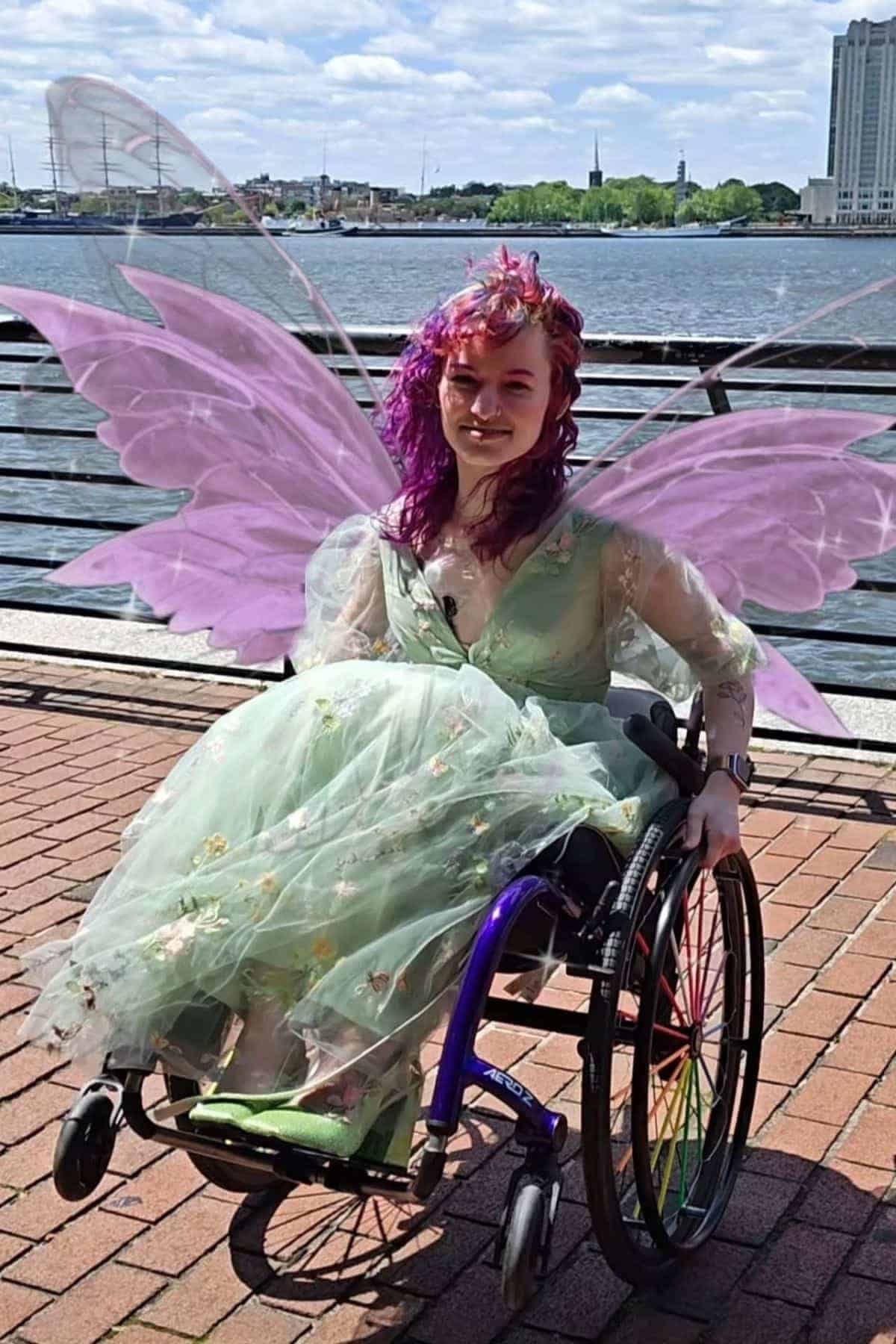 Person in a wheelchair wears a pastel green dress and fairy wings, sitting on a brick path by the waterfront with buildings and trees in the background.